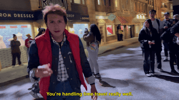 Person dressed like a 1980s movie character, wearing a red puffy vest and checkered shirt, walks toward the camera. Subtitles say: &quot;You're handling time travel really well.&quot;