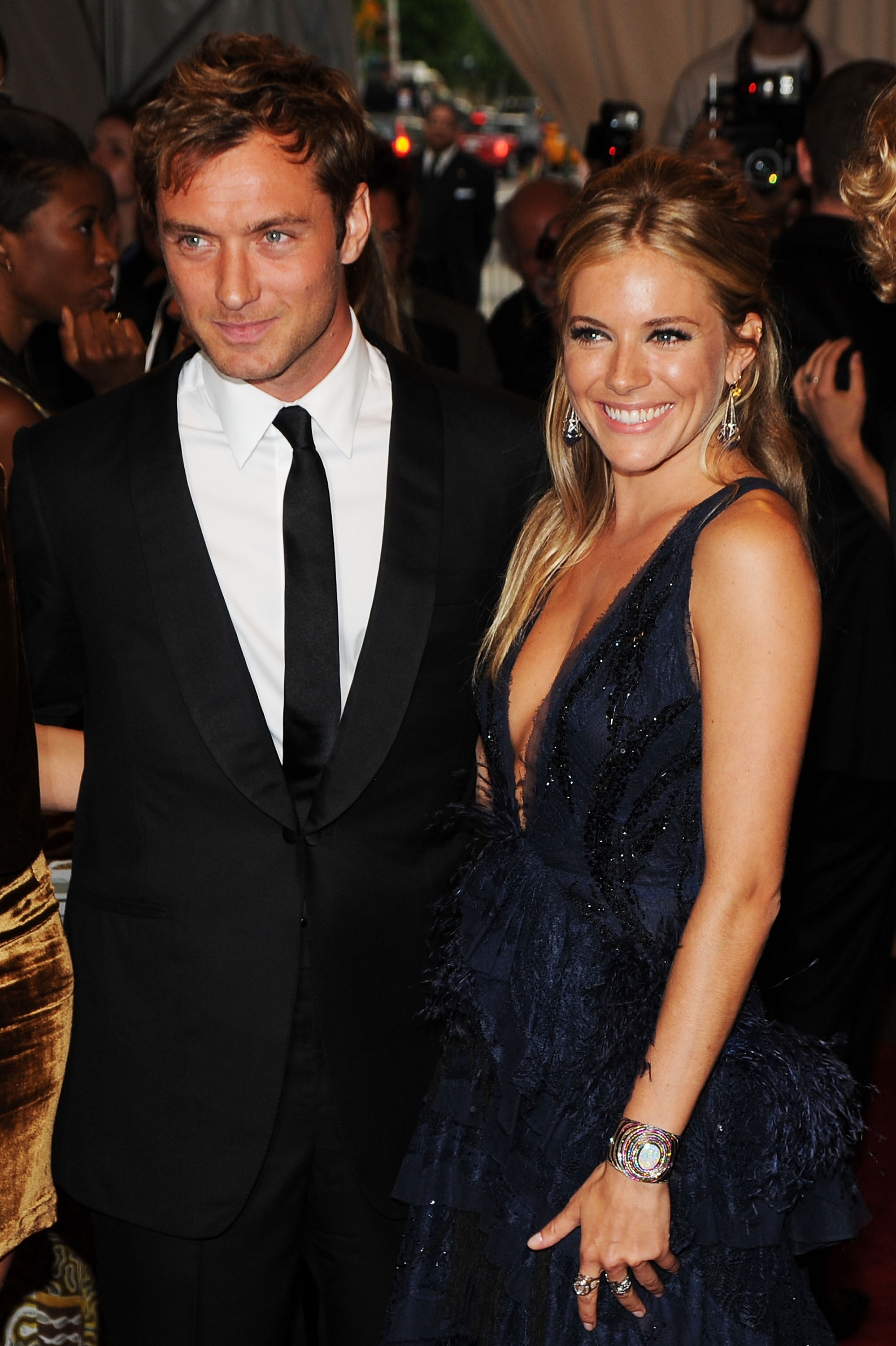 A man and woman on a red carpet. The woman is wearing an elegant deep V-neck gown with intricate detailing, and the man is in a classic suit and tie