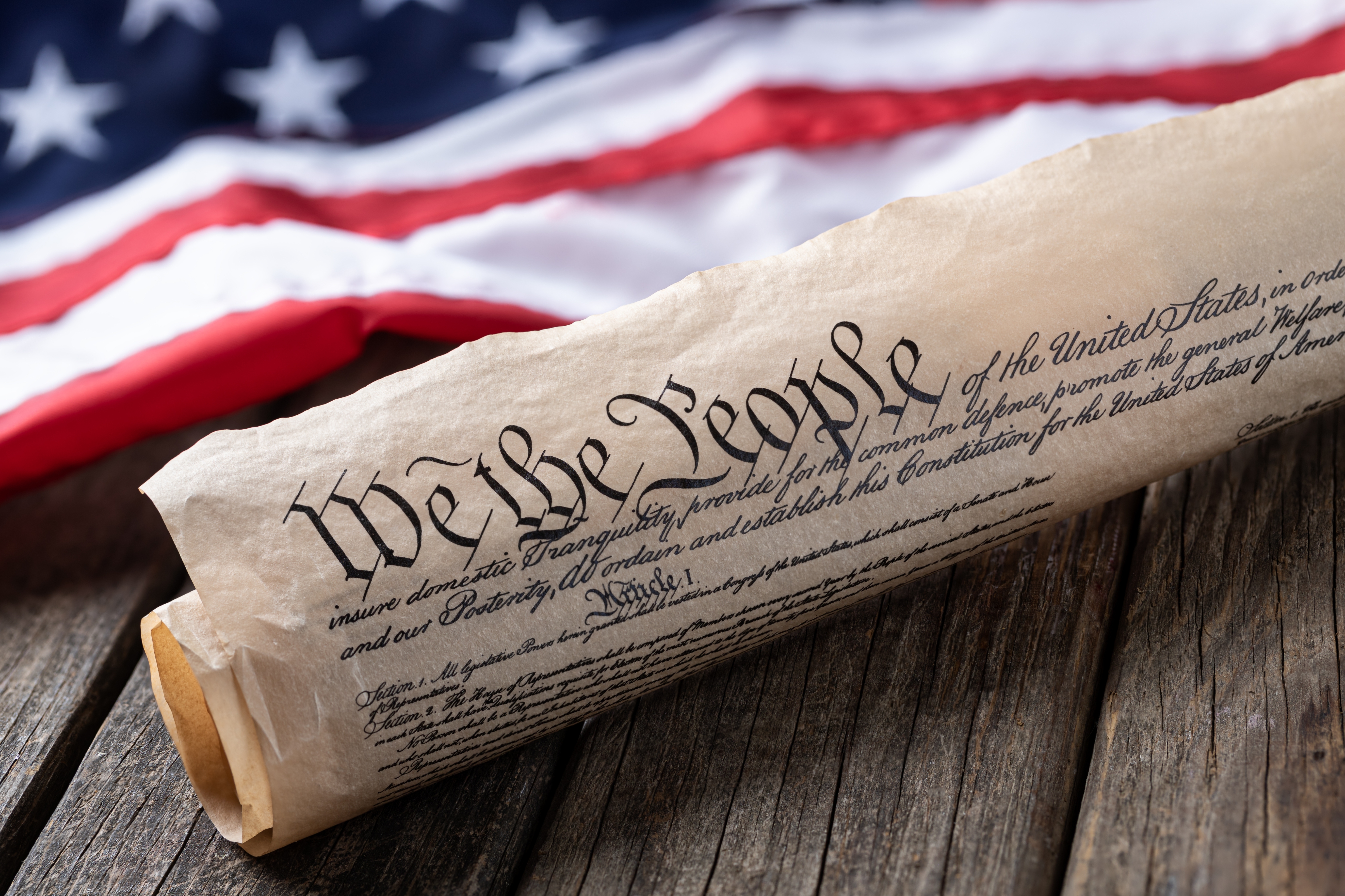 Rolled-up U.S. Constitution on wooden surface with an American flag in the background