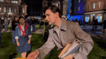 A person in vintage-style clothing holding papers outdoors, another person in period attire stands in the background amidst a crowd