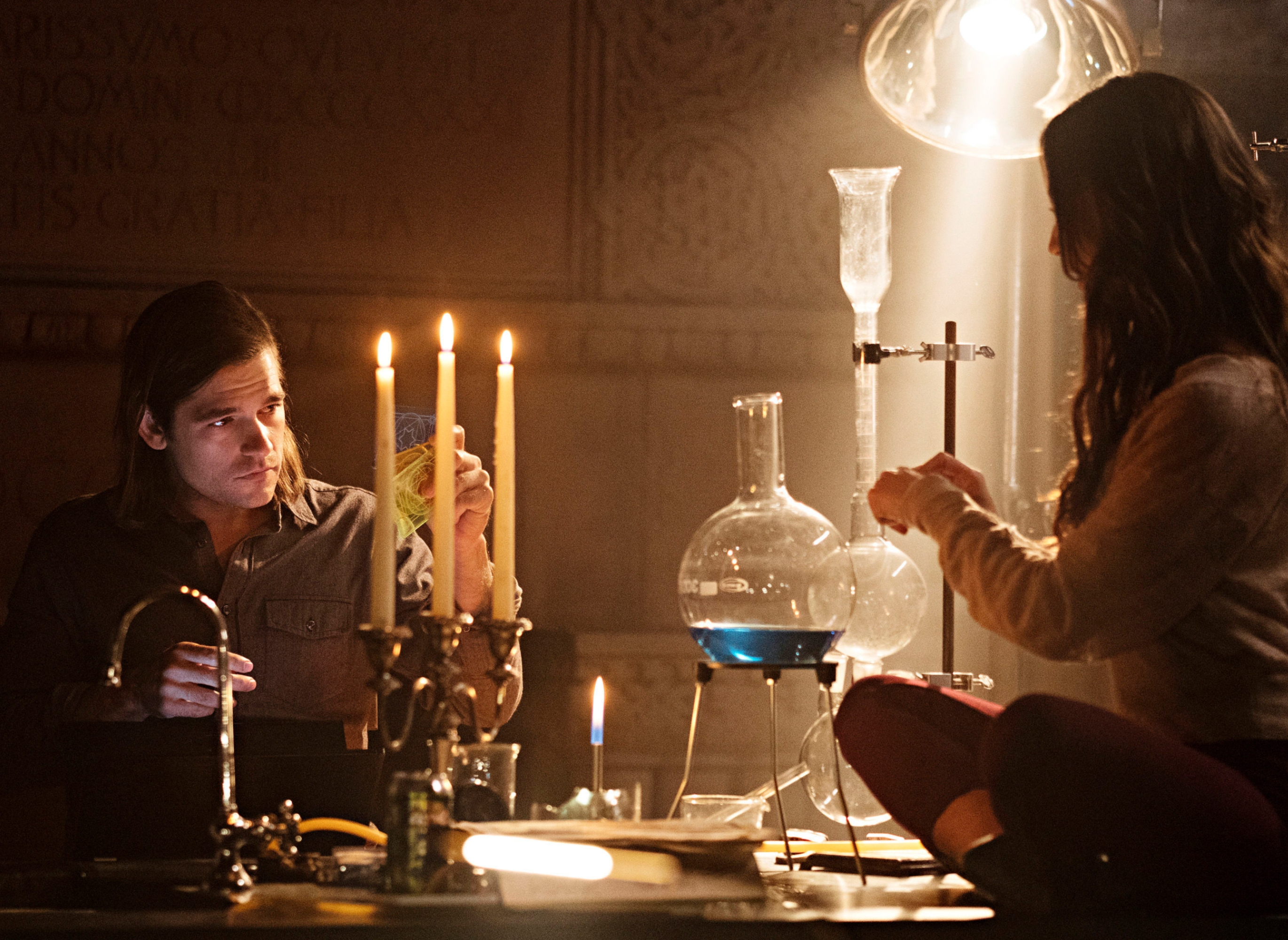 Two people perform a chemistry experiment in a dimly lit lab. One sits on a table, handling equipment; the other examines a flask under candlelight