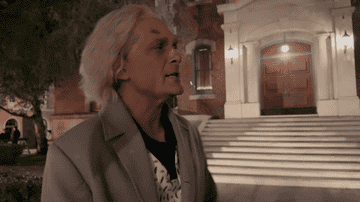 Person with white hair in a suit jacket stands outside a building with steps and large doors in the background