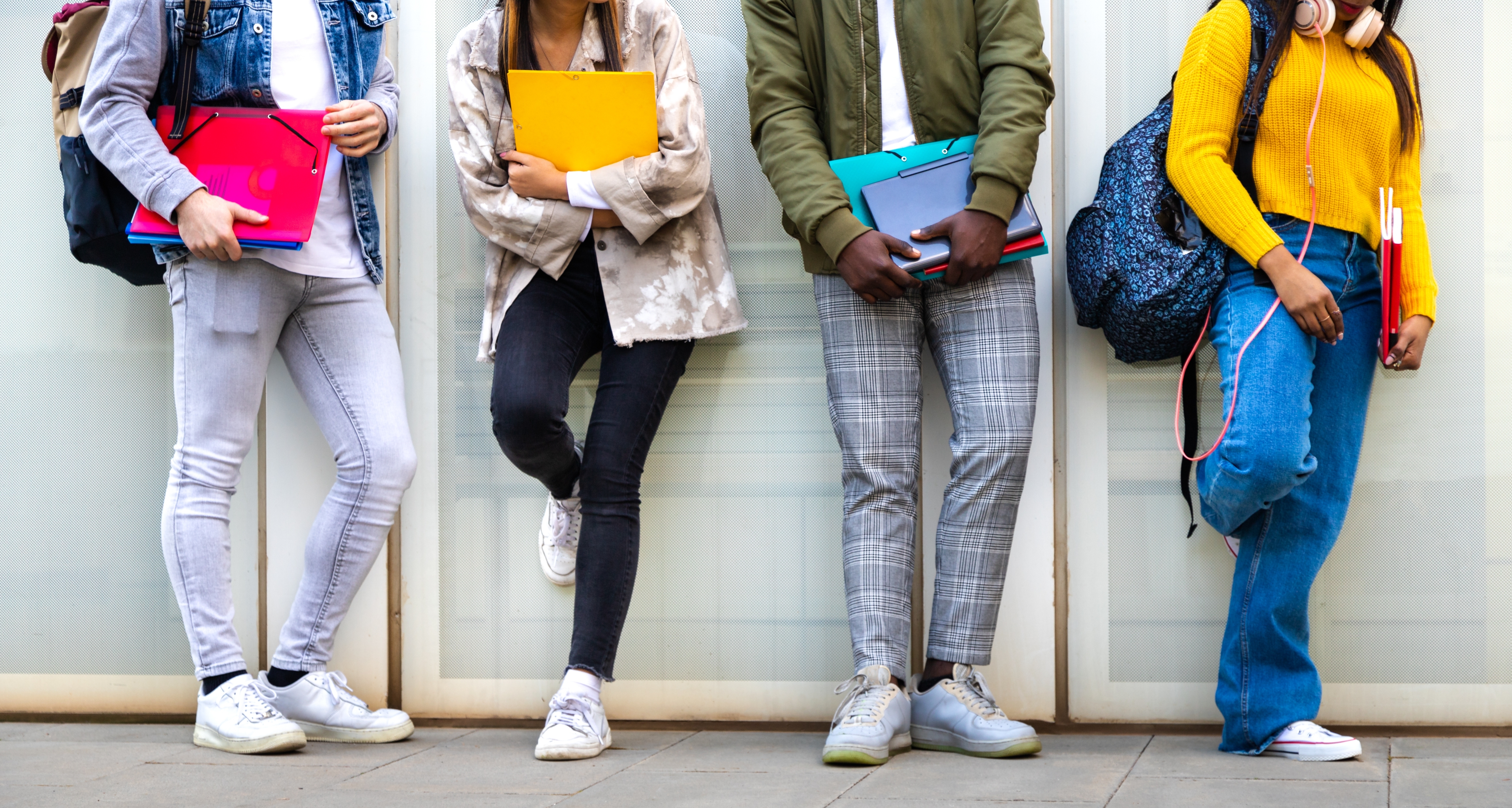 Four students stand against a wall, holding notebooks and wearing casual, trendy clothing, including jeans, jackets, and backpacks