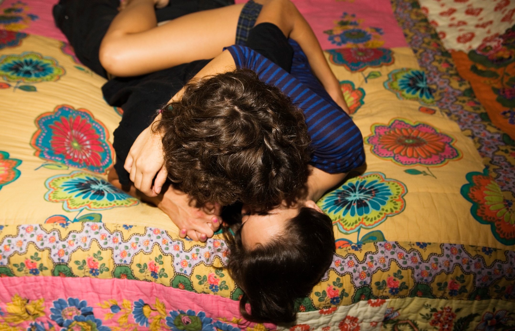 Two people, partially visible, lying close on a colorful patterned bedspread, with one person appearing to kiss the other's foot