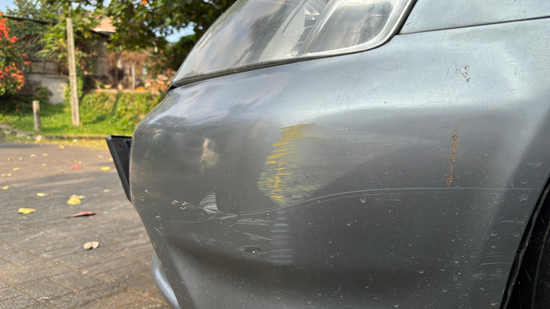 Close-up of a car with a dented bumper and scratch marks, parked on a residential street with trees in the background