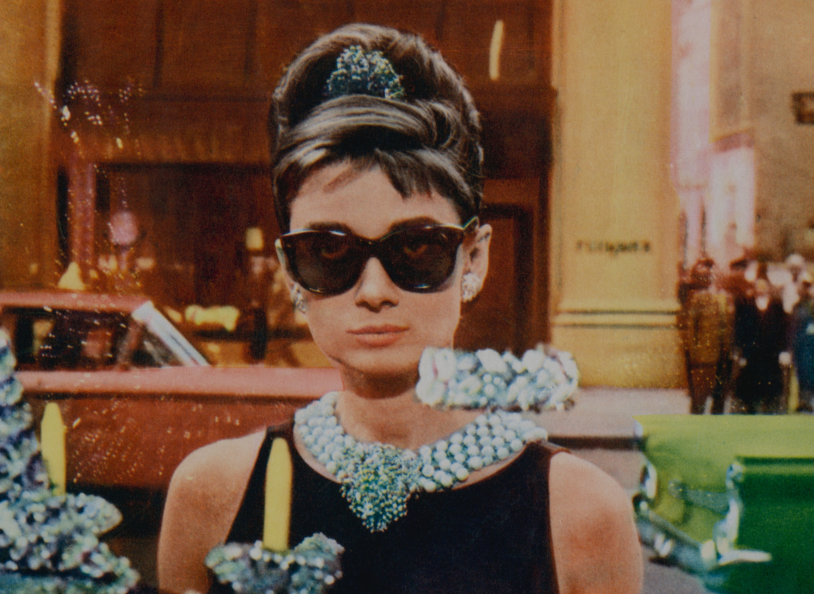 Person in elegant black dress, pearl necklace, and sunglasses holding a paper cup and bag, standing outdoors with cars and people in the background