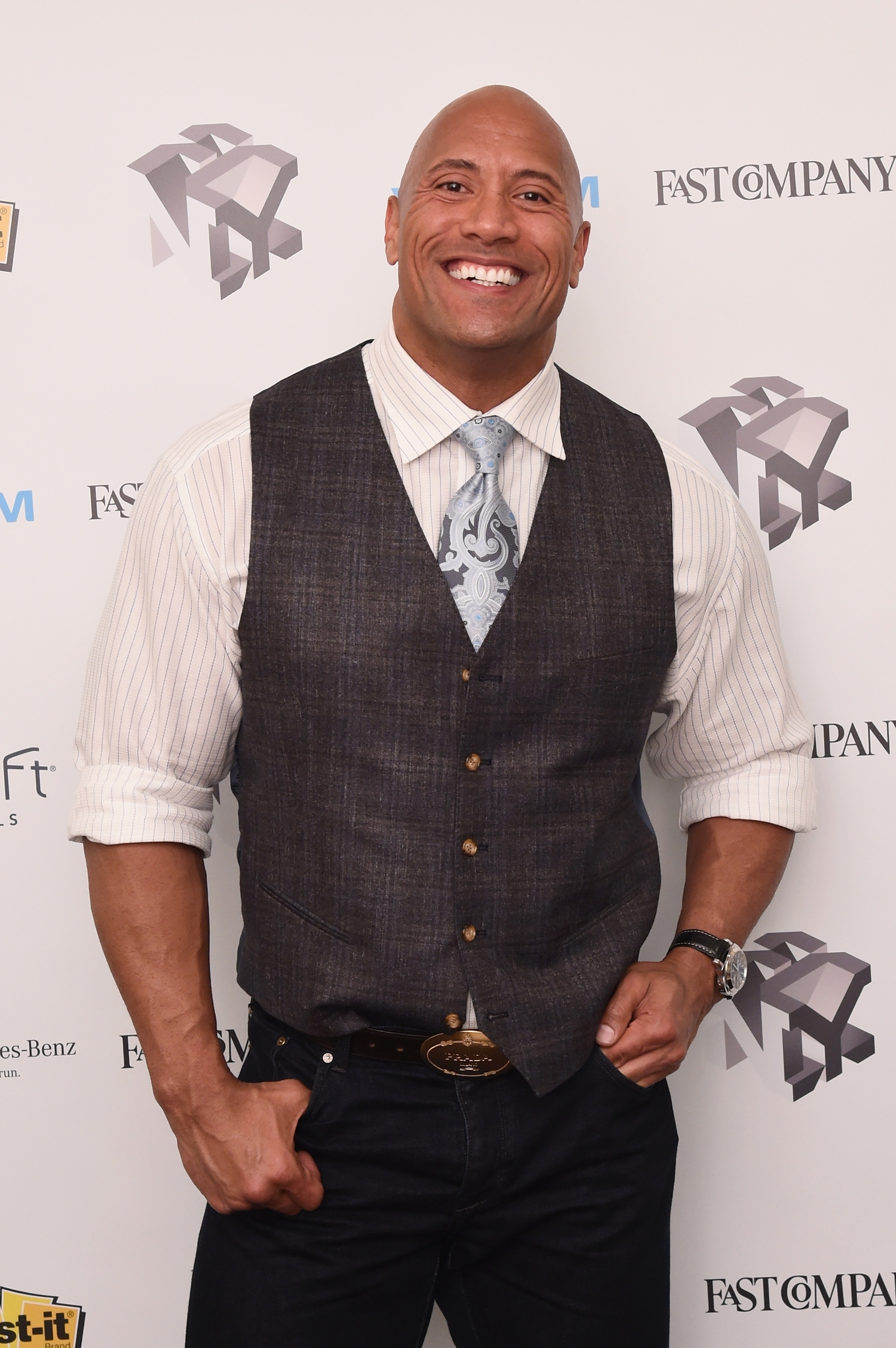 A smiling man in a dress shirt and a vest stands on a step-and-repeat backdrop for a celebrity event