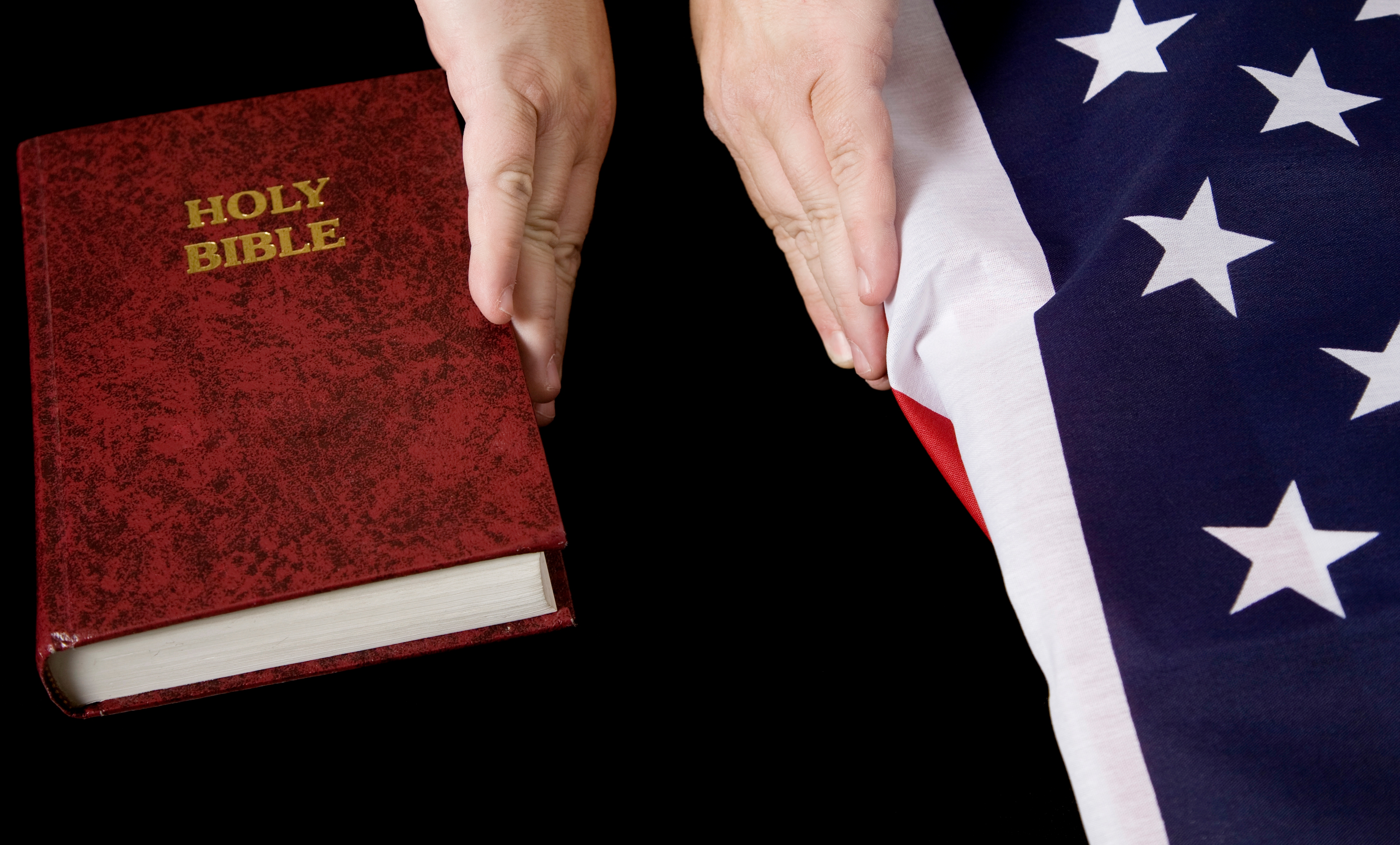 Hands hover over a Holy Bible next to an American flag on a black background
