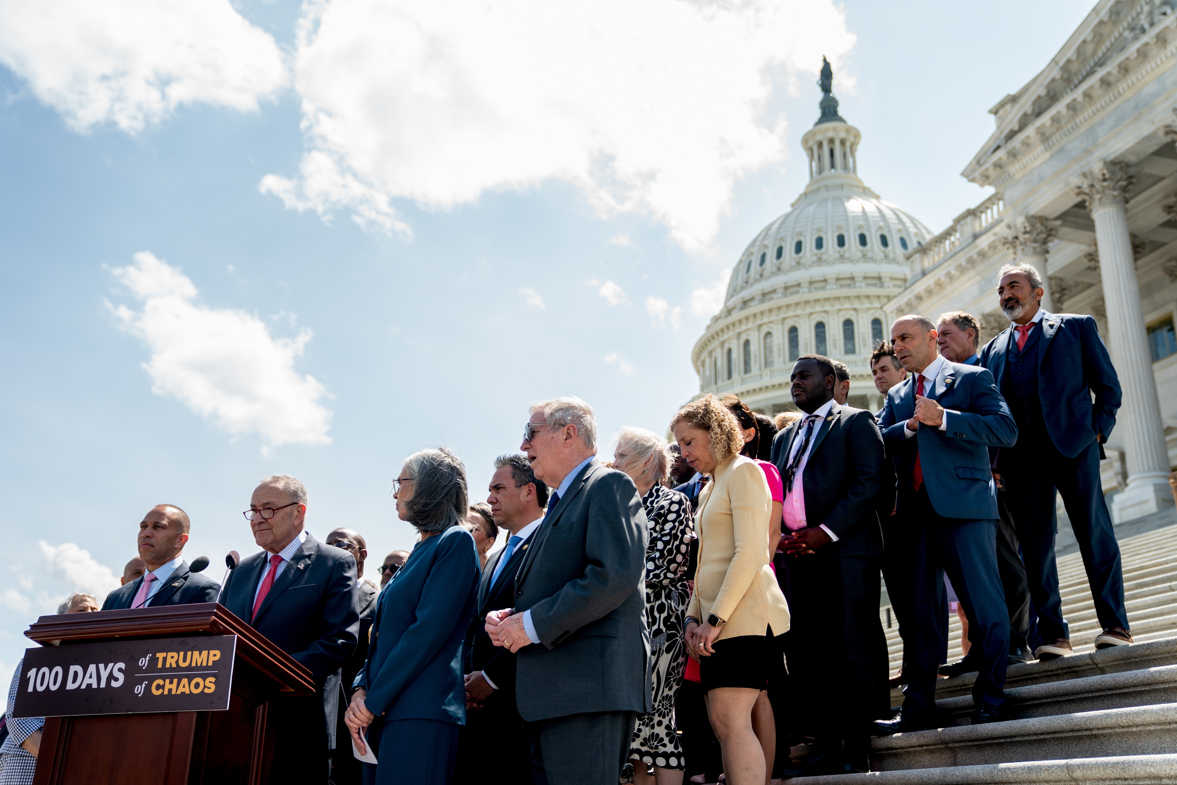 Group of people stand on U.S. Capitol steps during a press event. A sign reads "100 Days of Trump Chaos." Cloudy sky and Capitol dome in background