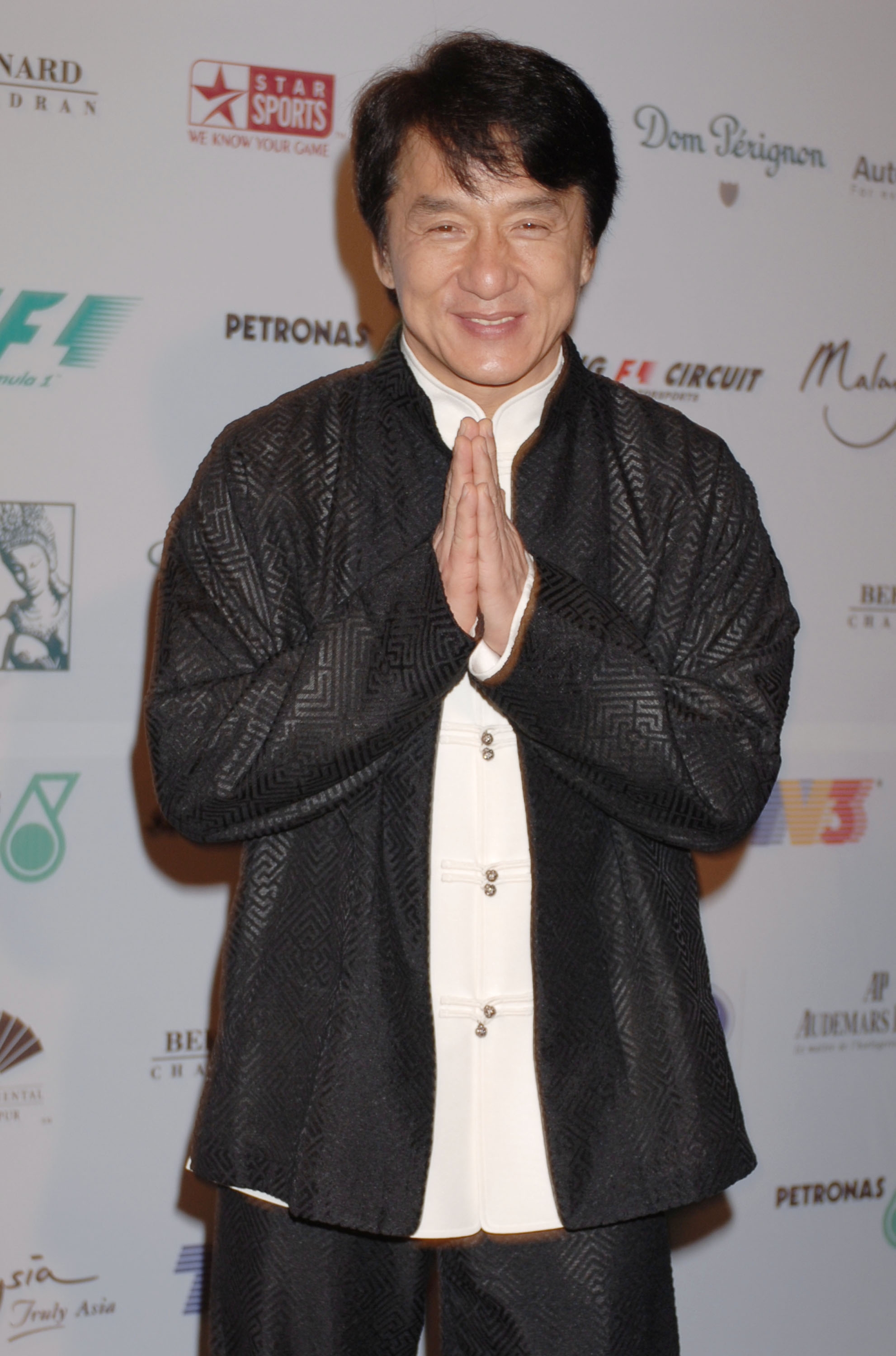 Person on the red carpet wearing a textured suit and traditional buttoned shirt, smiling with hands in a prayer gesture