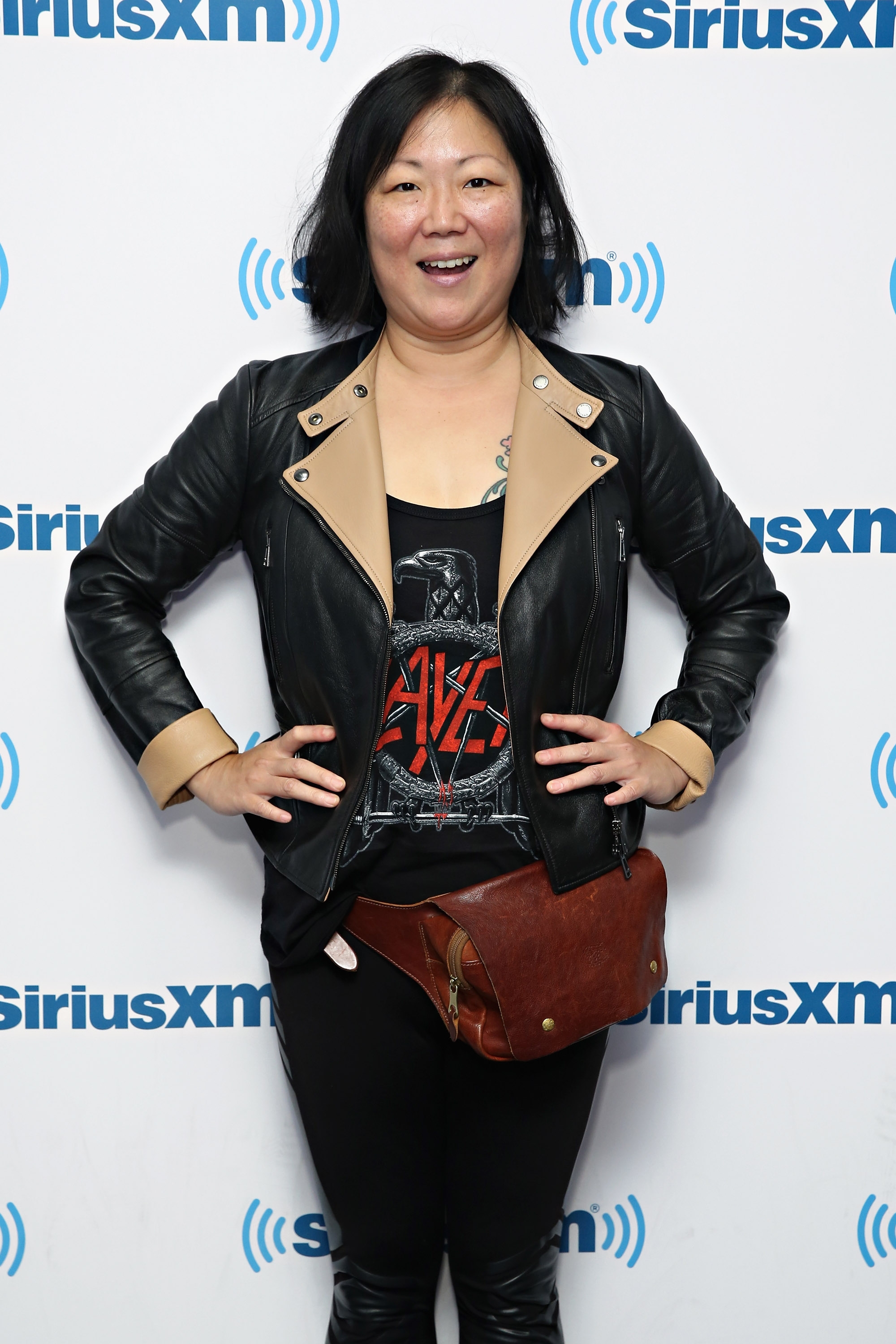 Person smiling at SiriusXM event, wearing a leather jacket and graphic tee, with a brown belt bag