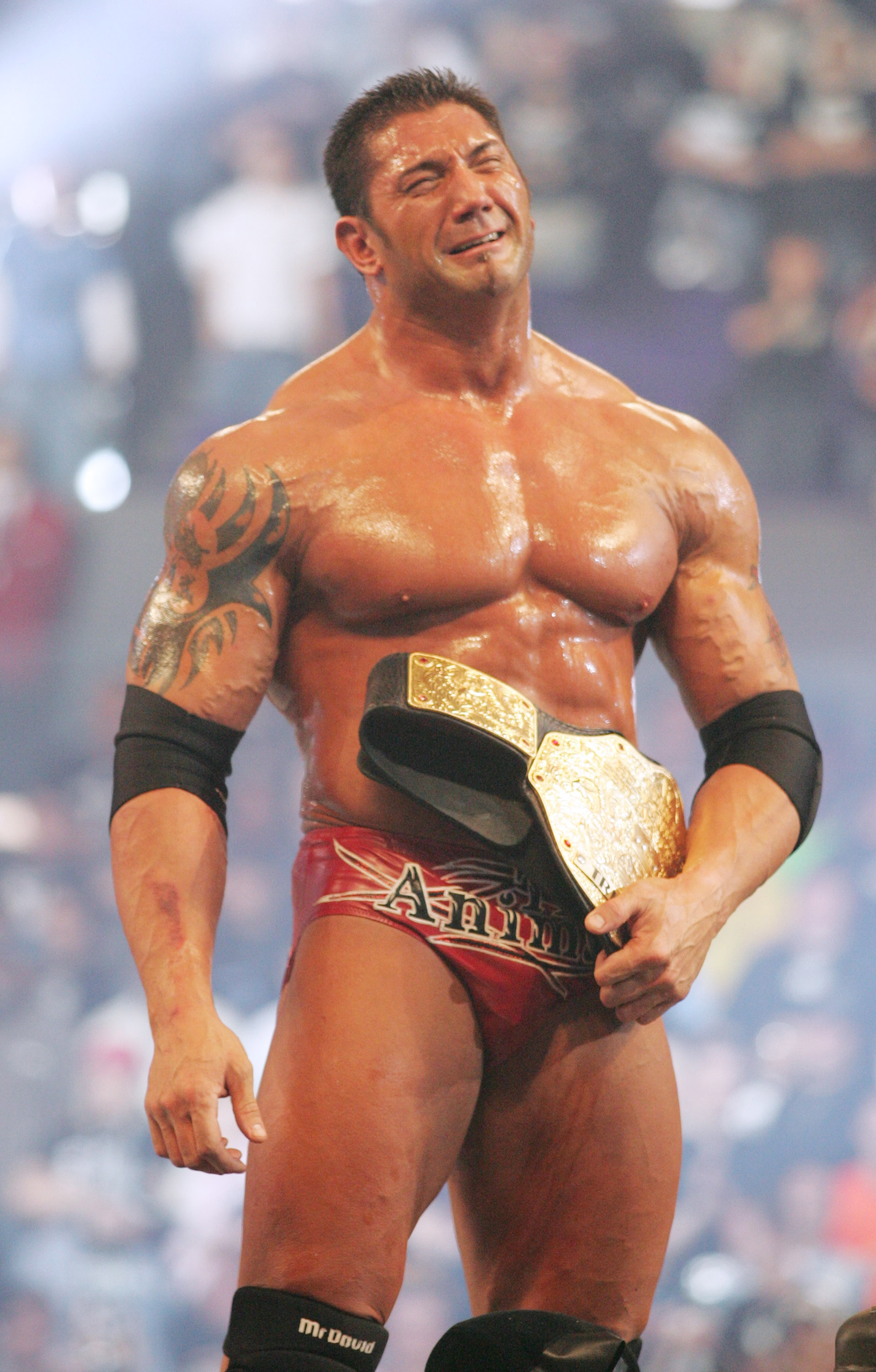 A muscular wrestler stands victoriously in a ring, holding a championship belt over his shoulder, wearing red trunks and black wristbands