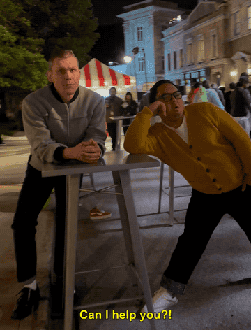 Two people at a high table, one in a casual jacket and the other in a yellow cardigan. Caption: &quot;Can I help you?!&quot;