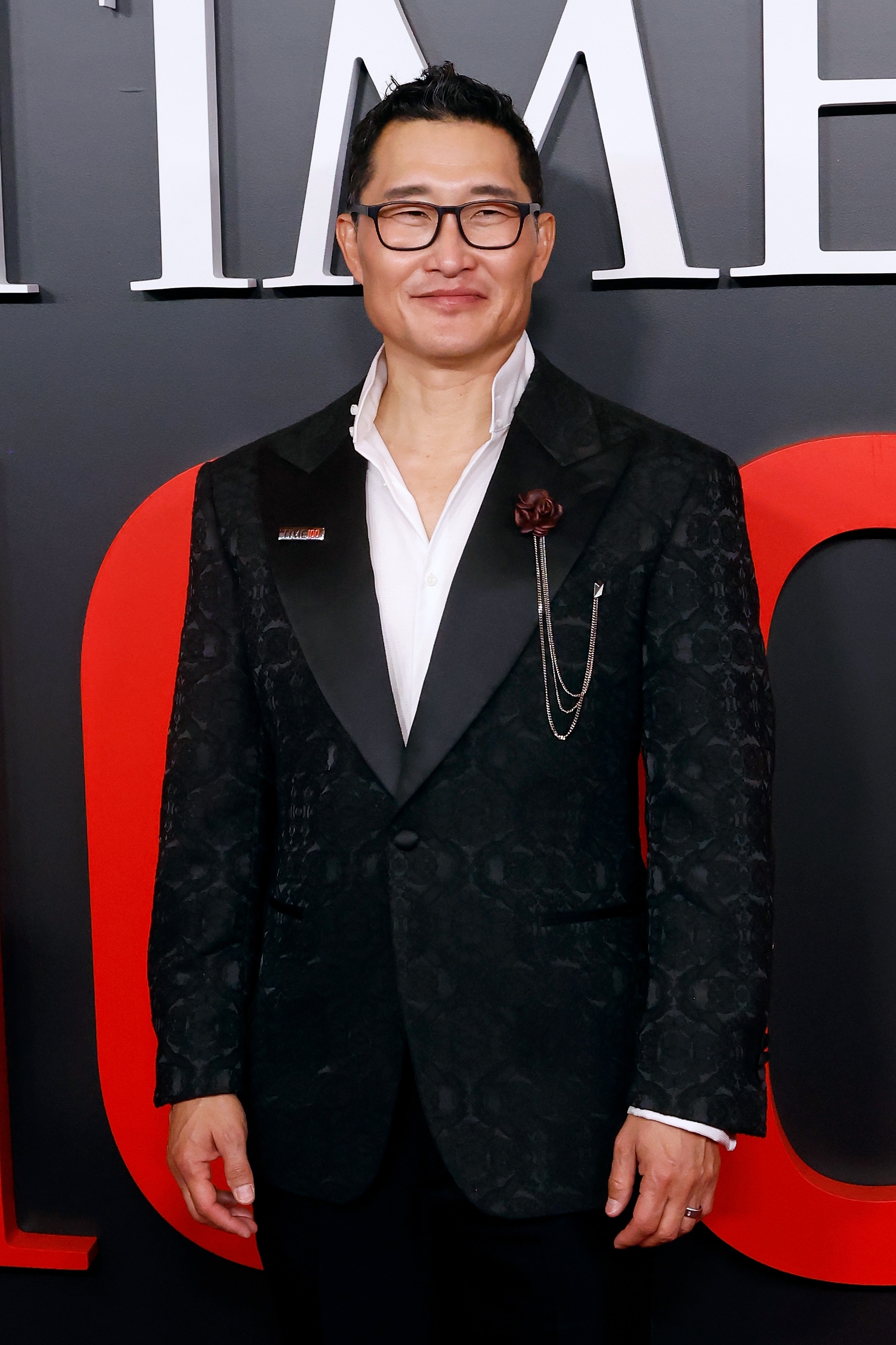 Person in an elegant black patterned suit with a white shirt and chain accessory, smiling on the red carpet