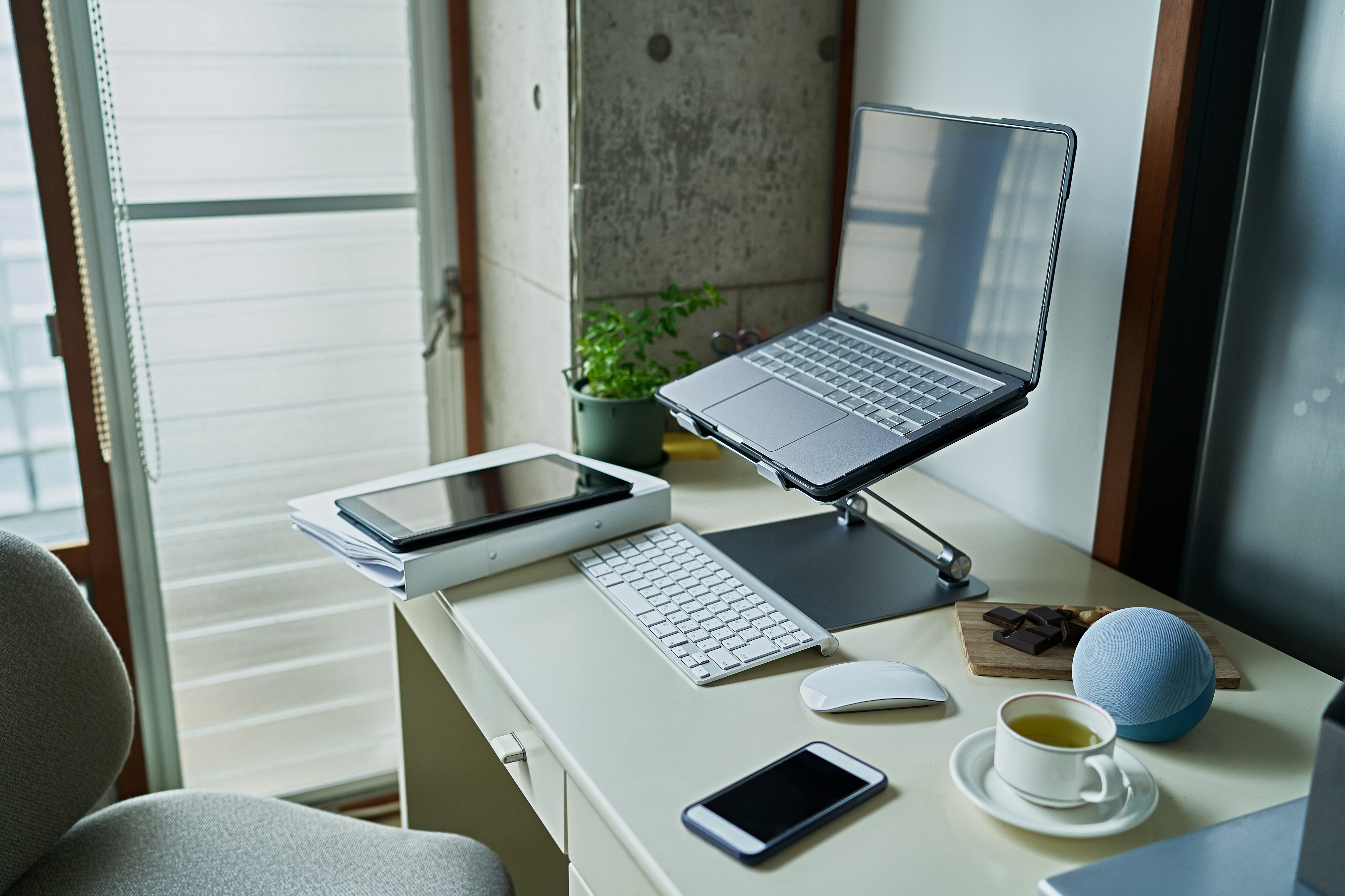 Desk setup with a laptop on a stand, keyboard, tablet, smartphone, mouse, cup of tea, speaker, and potted plant by a window