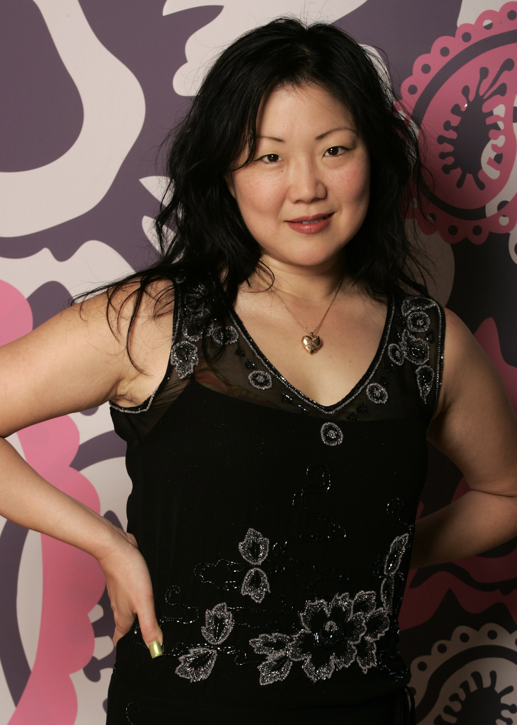 Person in floral-patterned dress and heart necklace stands confidently against a bold, abstract background