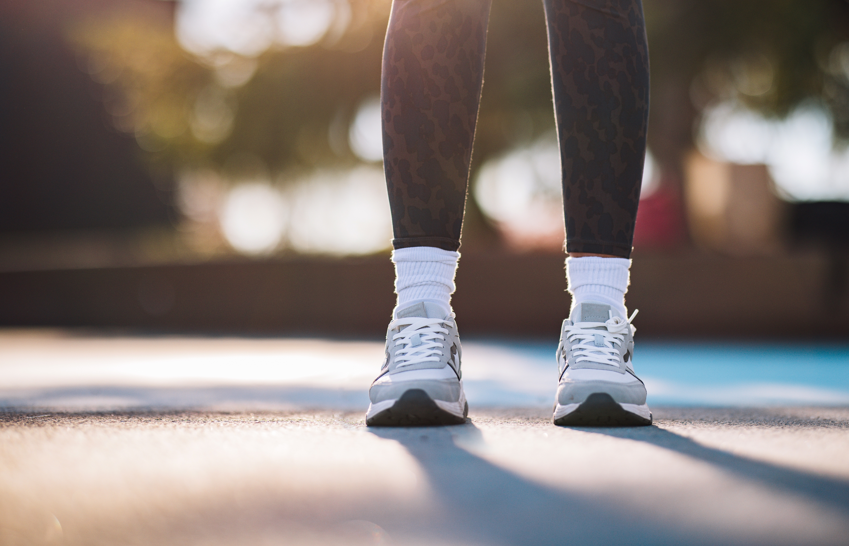 A person wearing athletic shoes, leggings, and white socks stands on a sunny outdoor surface, hinting at a casual or sporty setting