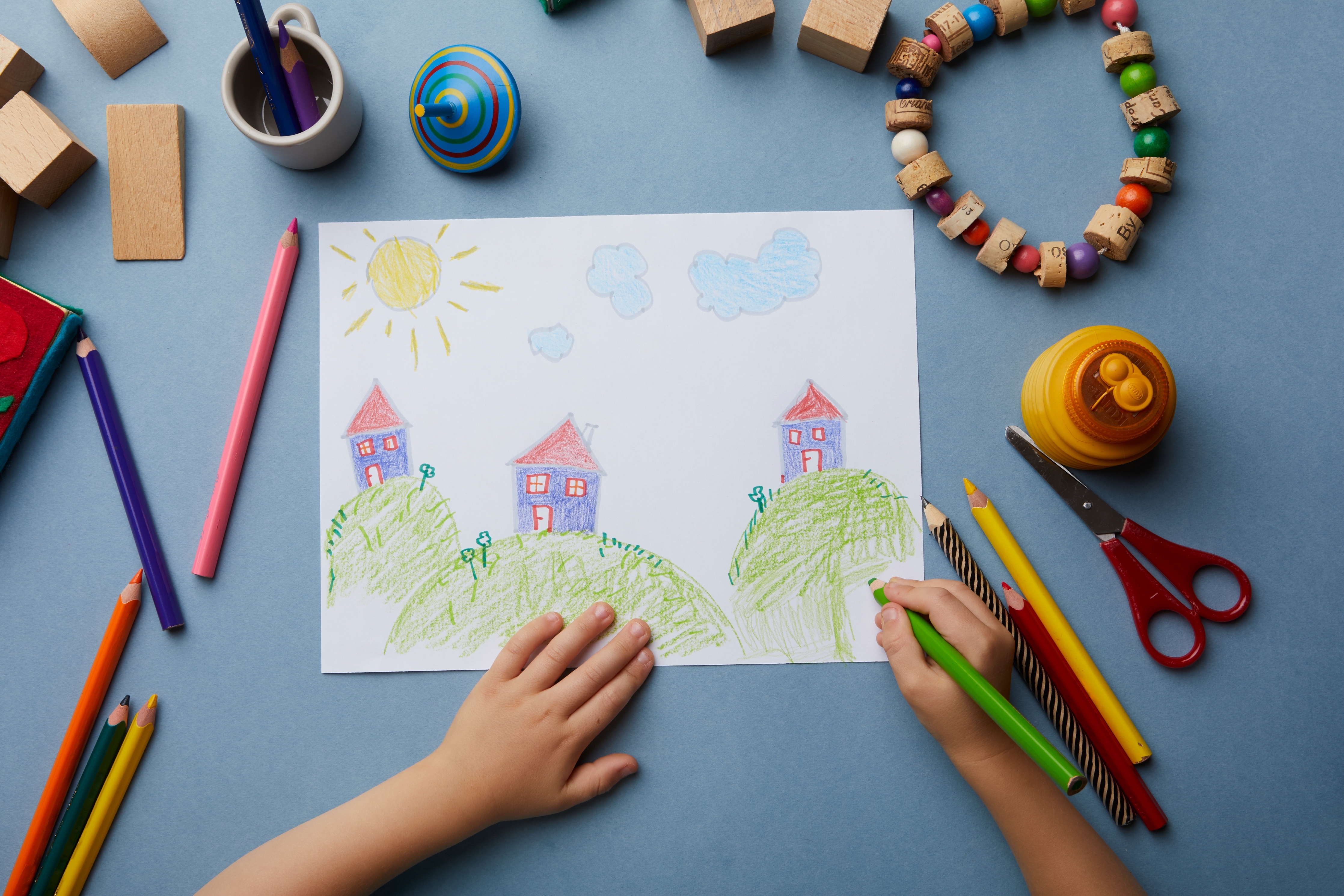 Child drawing a cheerful landscape with houses, hills, sun, and clouds, surrounded by crayons and toys on a table