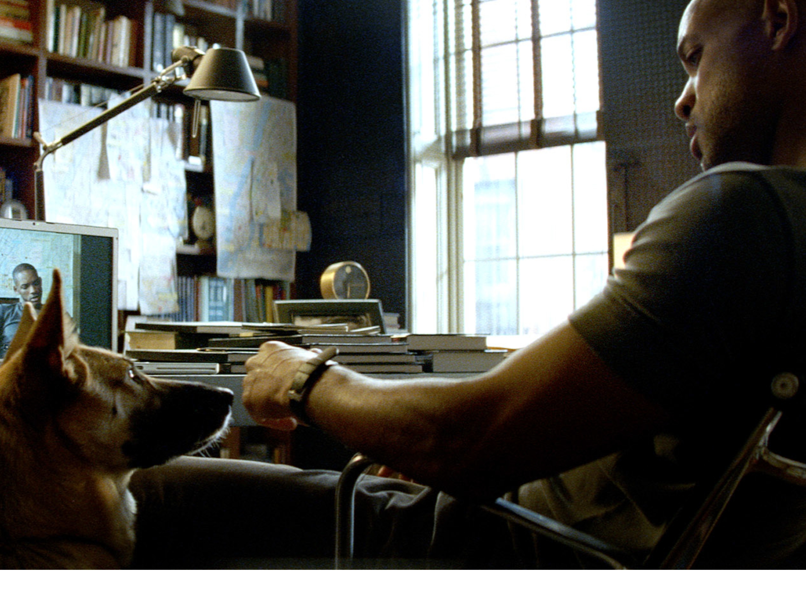 Person sits at a desk with a laptop, looking down at a dog. The setting is a study with books and papers on the walls