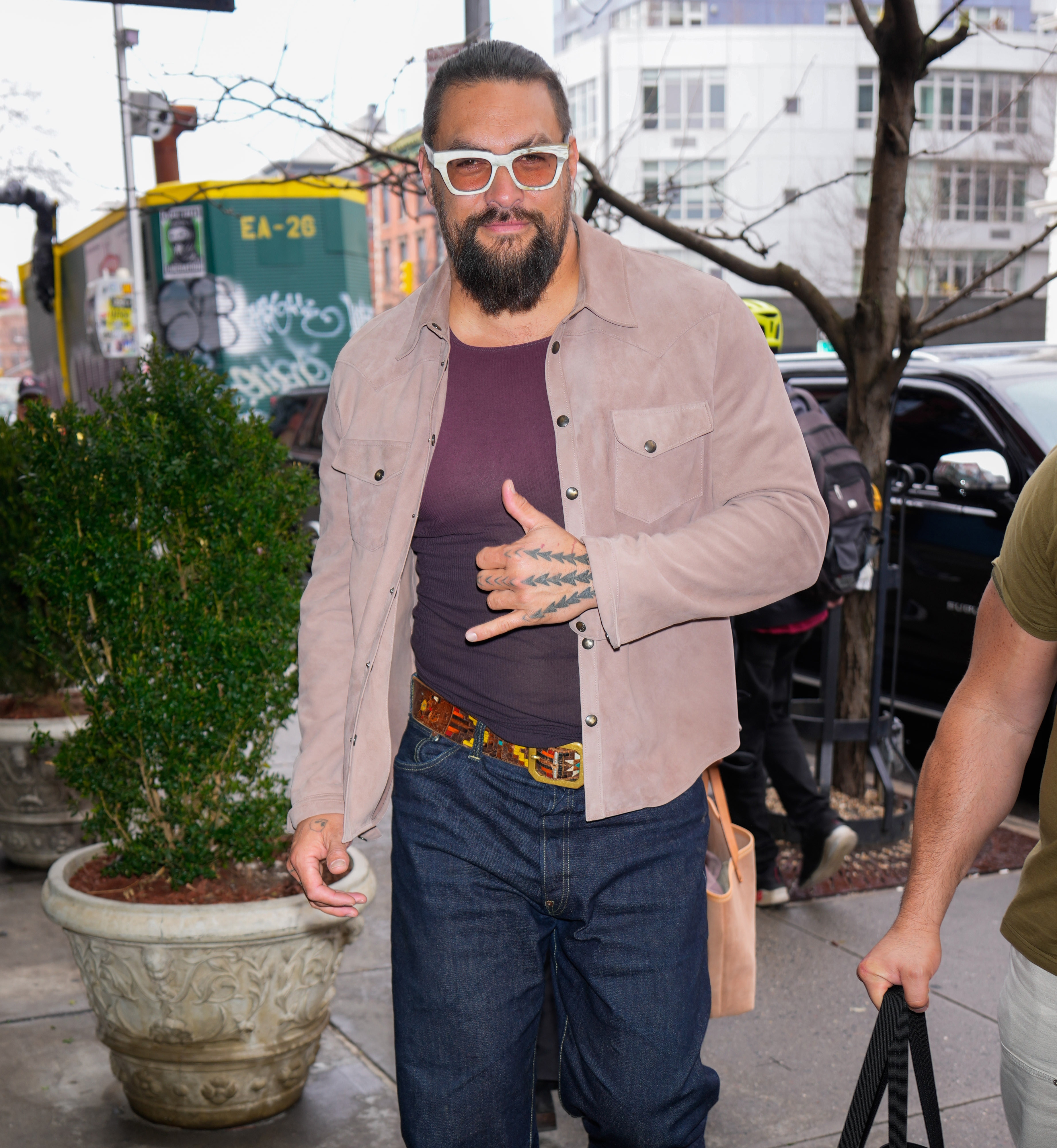 Person wearing casual suede jacket and jeans, with sunglasses and a beard, making a friendly gesture while walking outdoors
