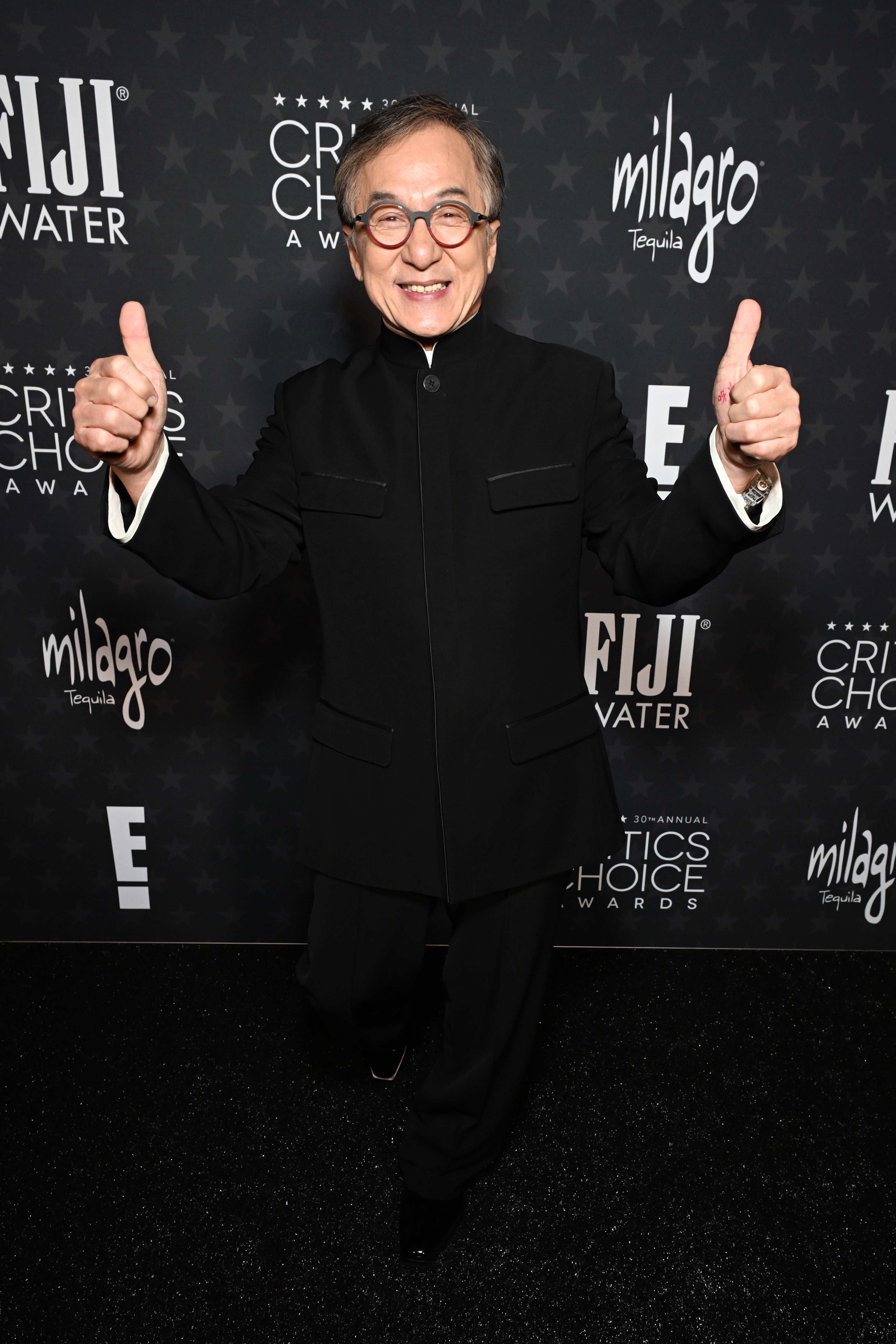 Person in a dark suit on the Critics Choice Awards carpet, giving two thumbs up, smiling with glasses