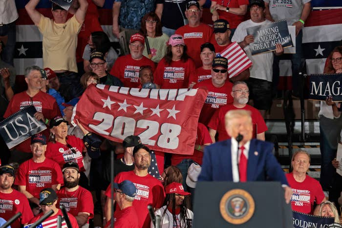 Crowd at a rally holding "Trump 2028" banner and flags, with many wearing hats and shirts supporting a political figure