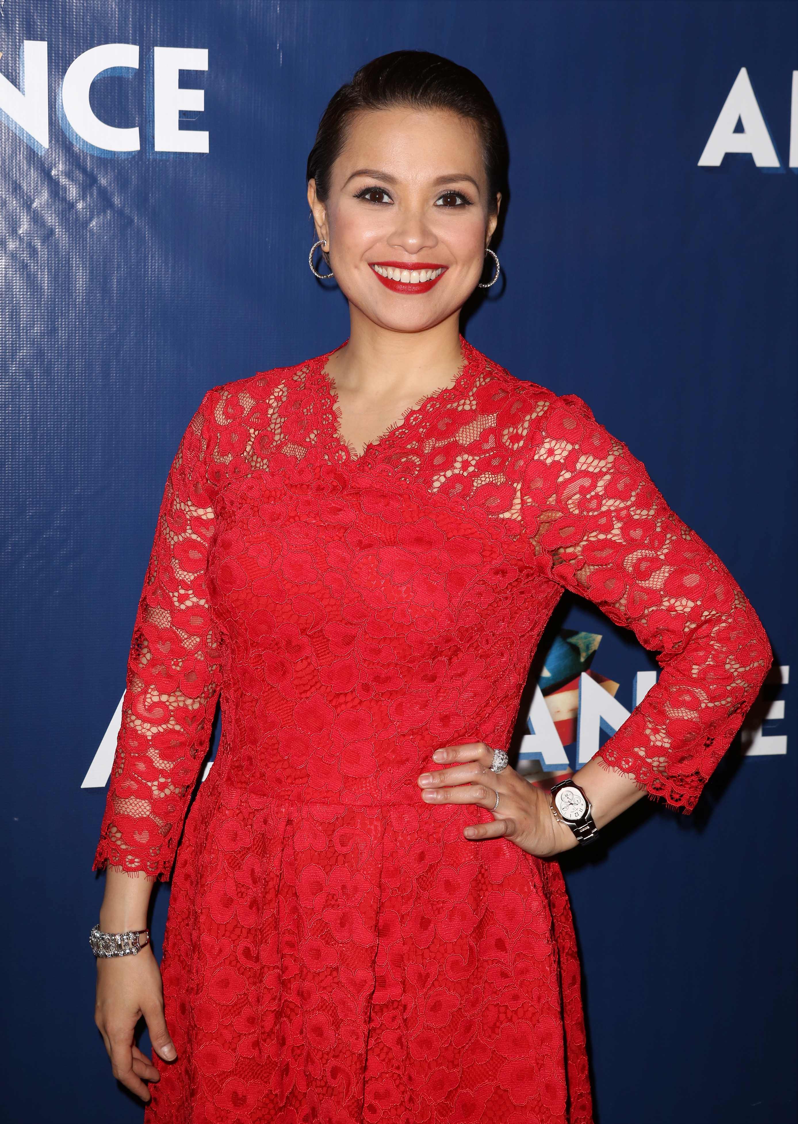 Celebrity in a stylish lace dress, posing on the red carpet with a smile, hands on hips, against a backdrop with partial text visible