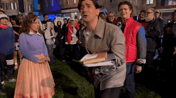 A person in a casual jacket holds a stack of books, while another in a pink skirt and cardigan looks concerned. A crowd watches in a lively setting