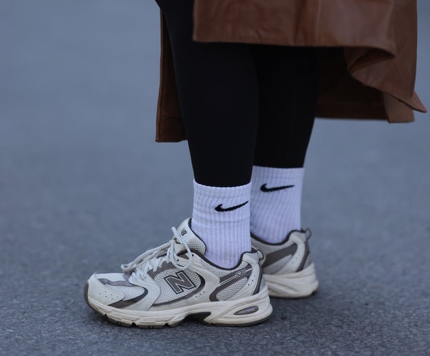Person wearing black leggings, white Nike socks, and white New Balance sneakers. Brown coat hem visible. Casual style