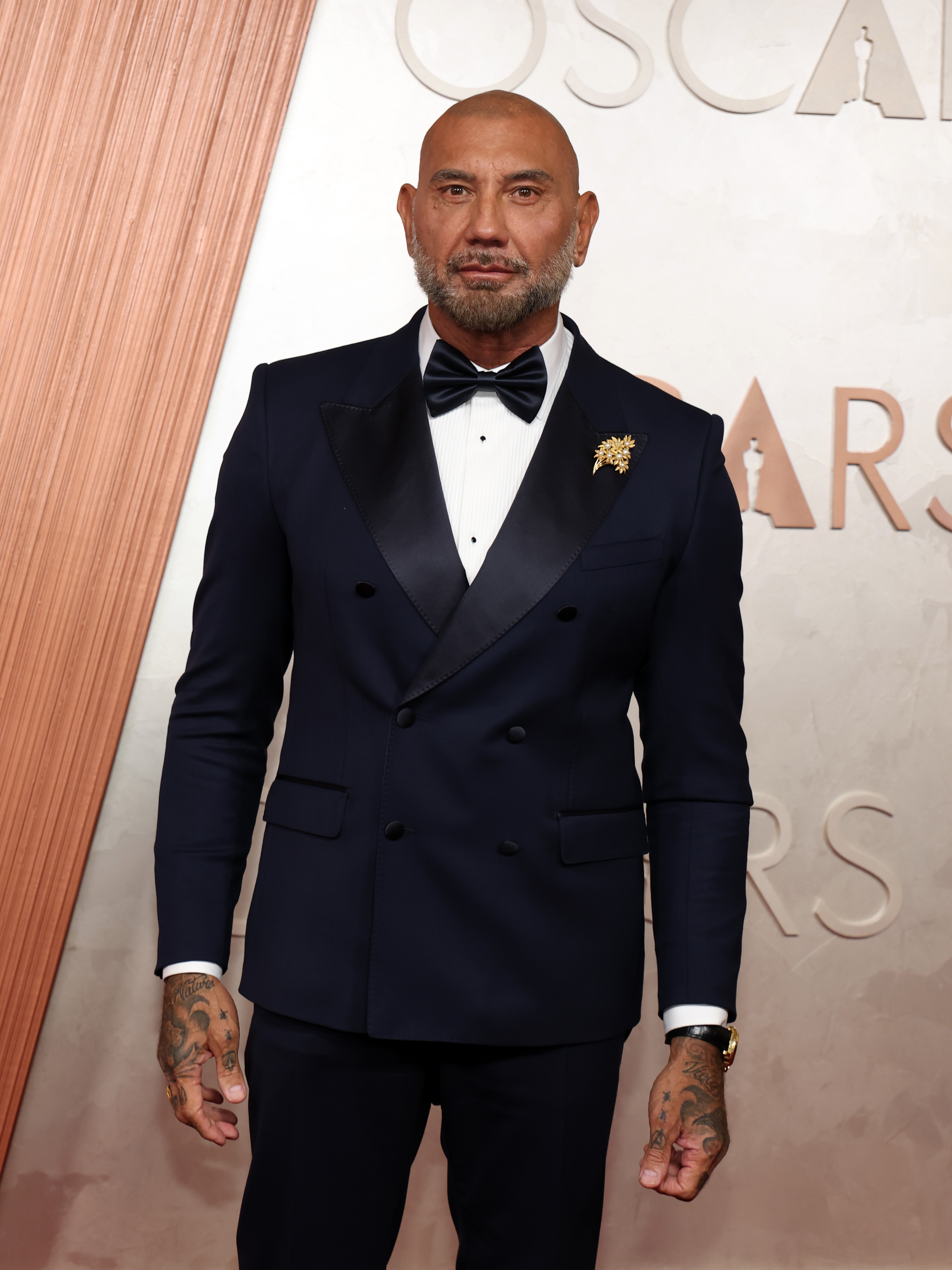 Person in a tailored tuxedo with a bow tie and a lapel pin, posing on the red carpet at an event