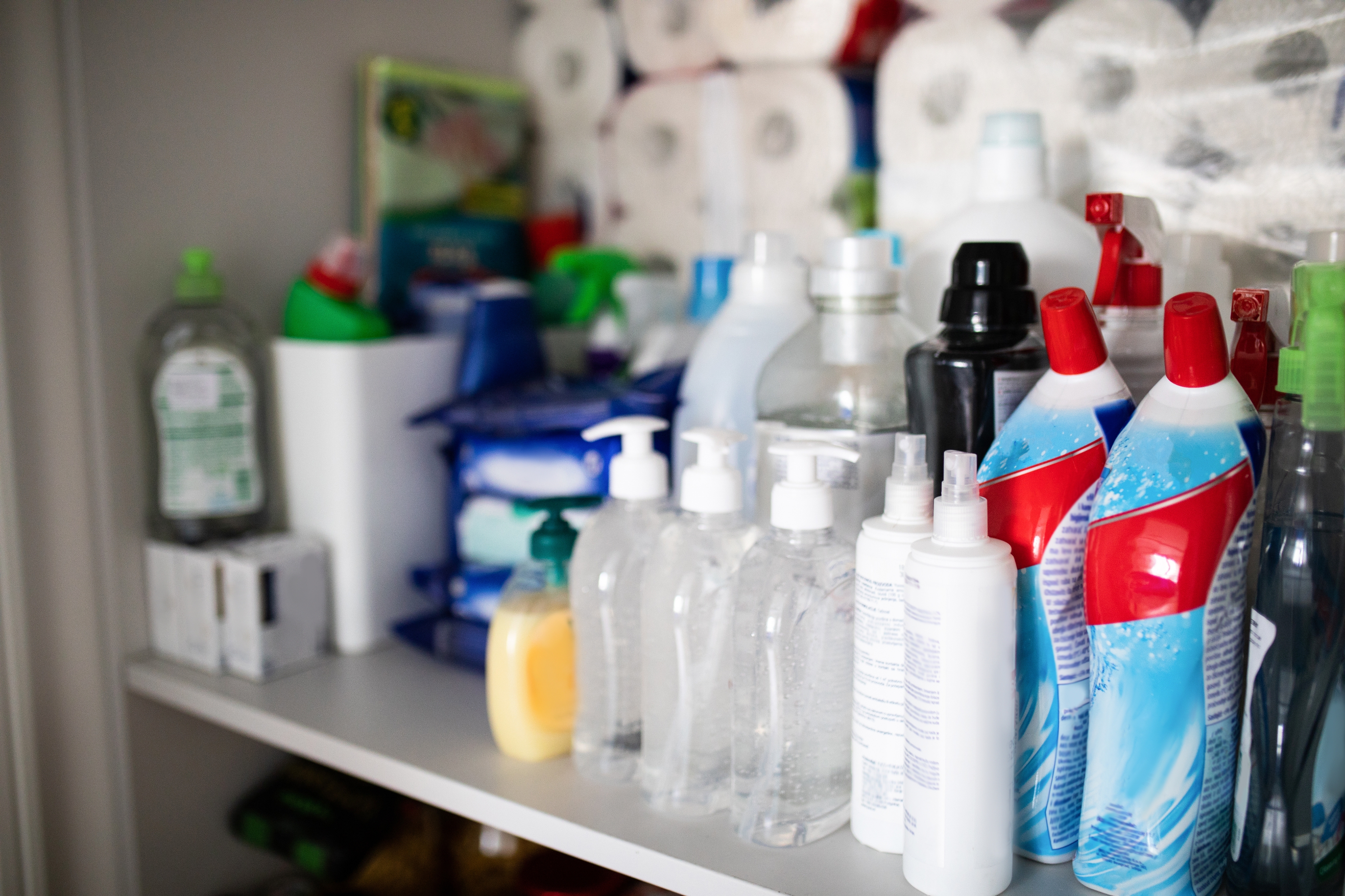 Various cleaning supplies and hygiene products are organized on a shelf, including bottles, sprays, packs, and rolls of paper towels