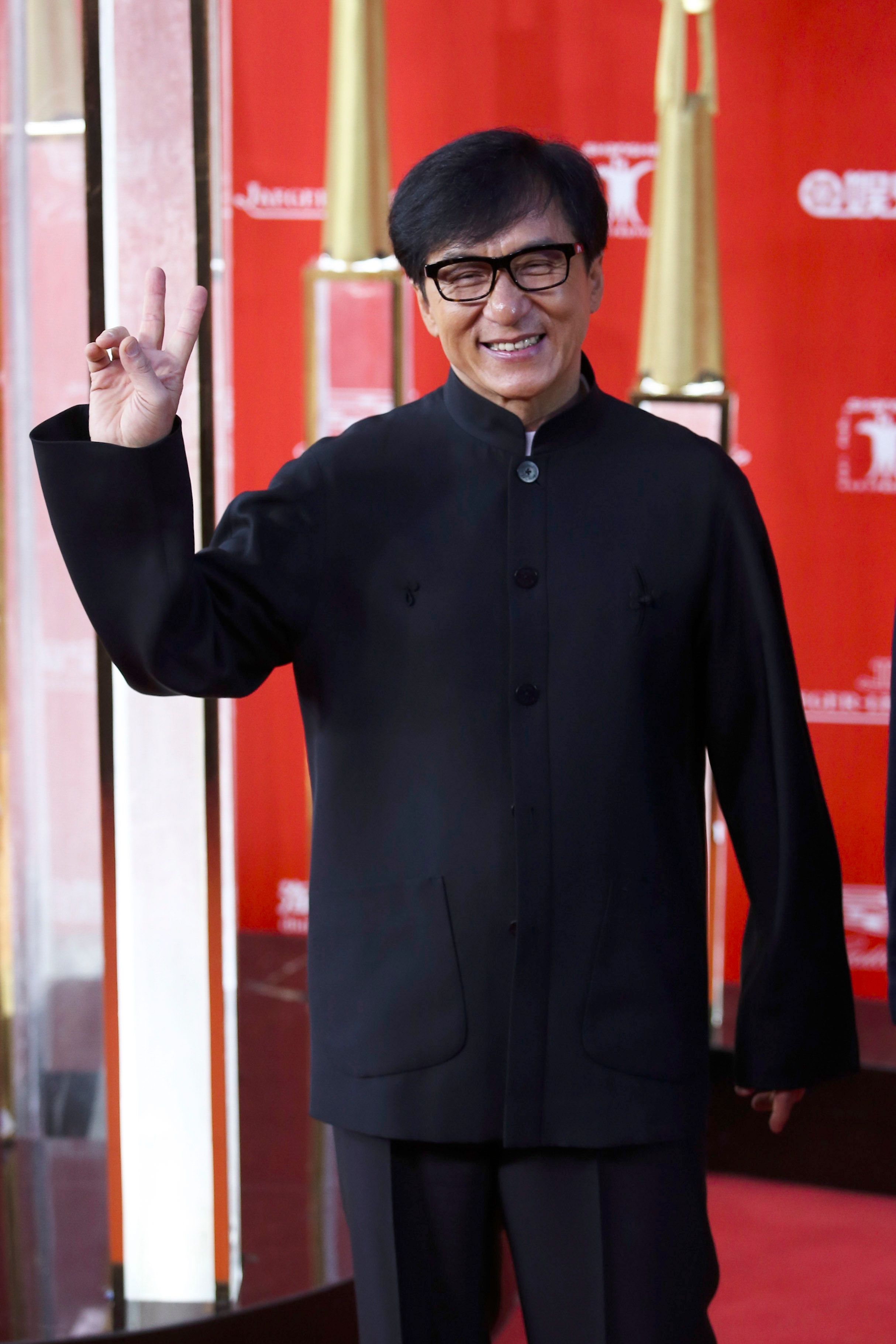 Individual in traditional black attire on red carpet, smiling and making a peace sign gesture