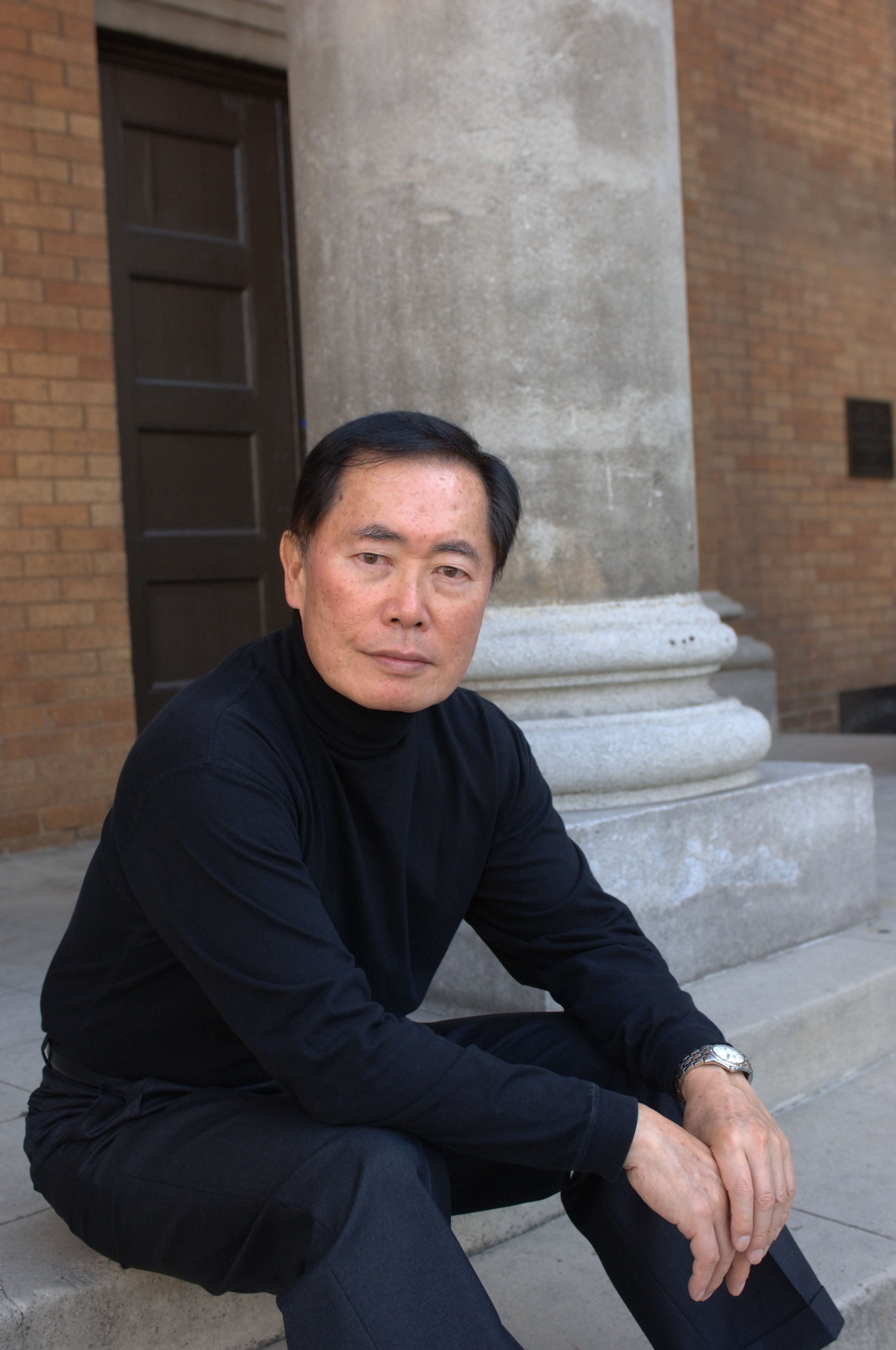 Individual sitting on steps with a serious expression, wearing a sleek, modern black turtleneck and matching pants