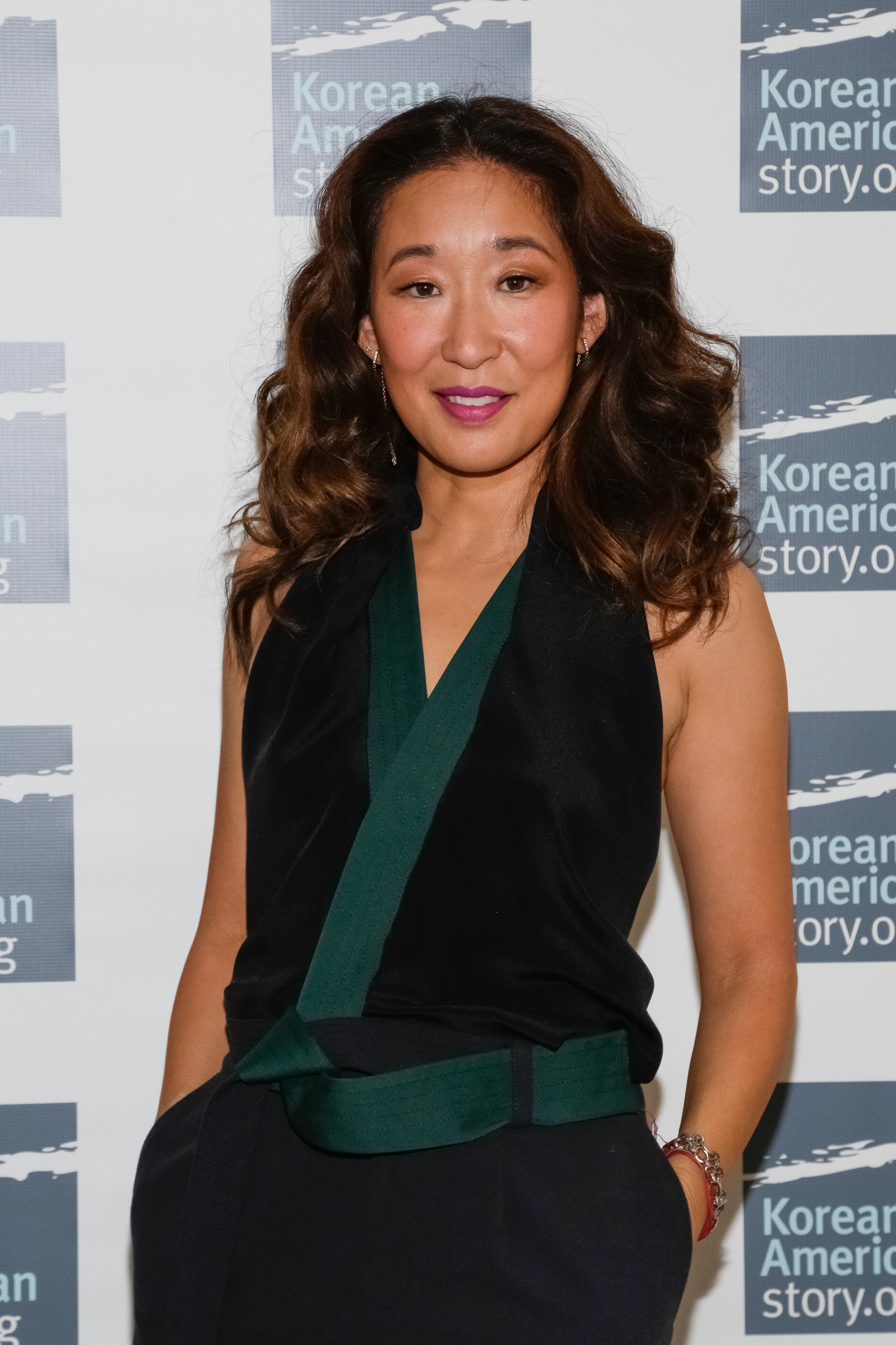 Person in stylish black and green outfit at an event in front of KoreanAmericanStory.org backdrop, smiling confidently