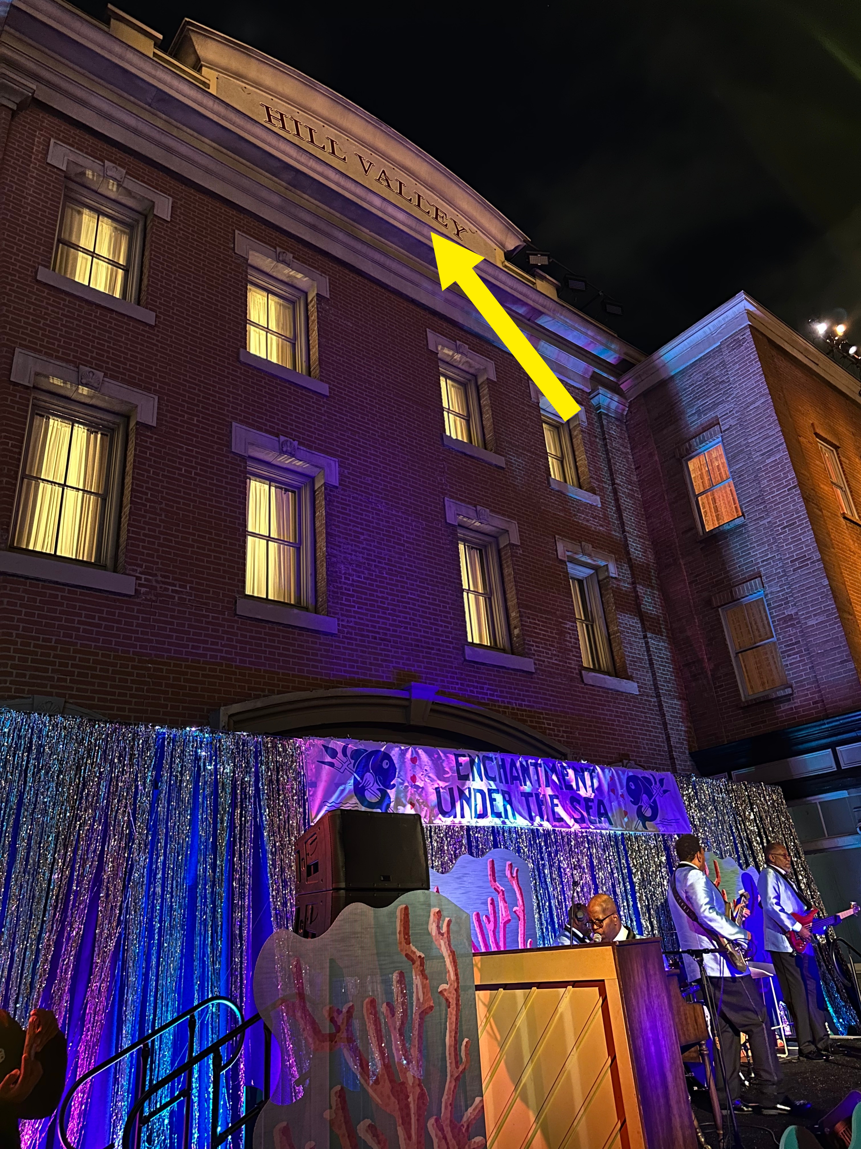 Building exterior at night with &quot;Hill Valley&quot; sign. Stage below with glittery backdrop and performers under colorful lights