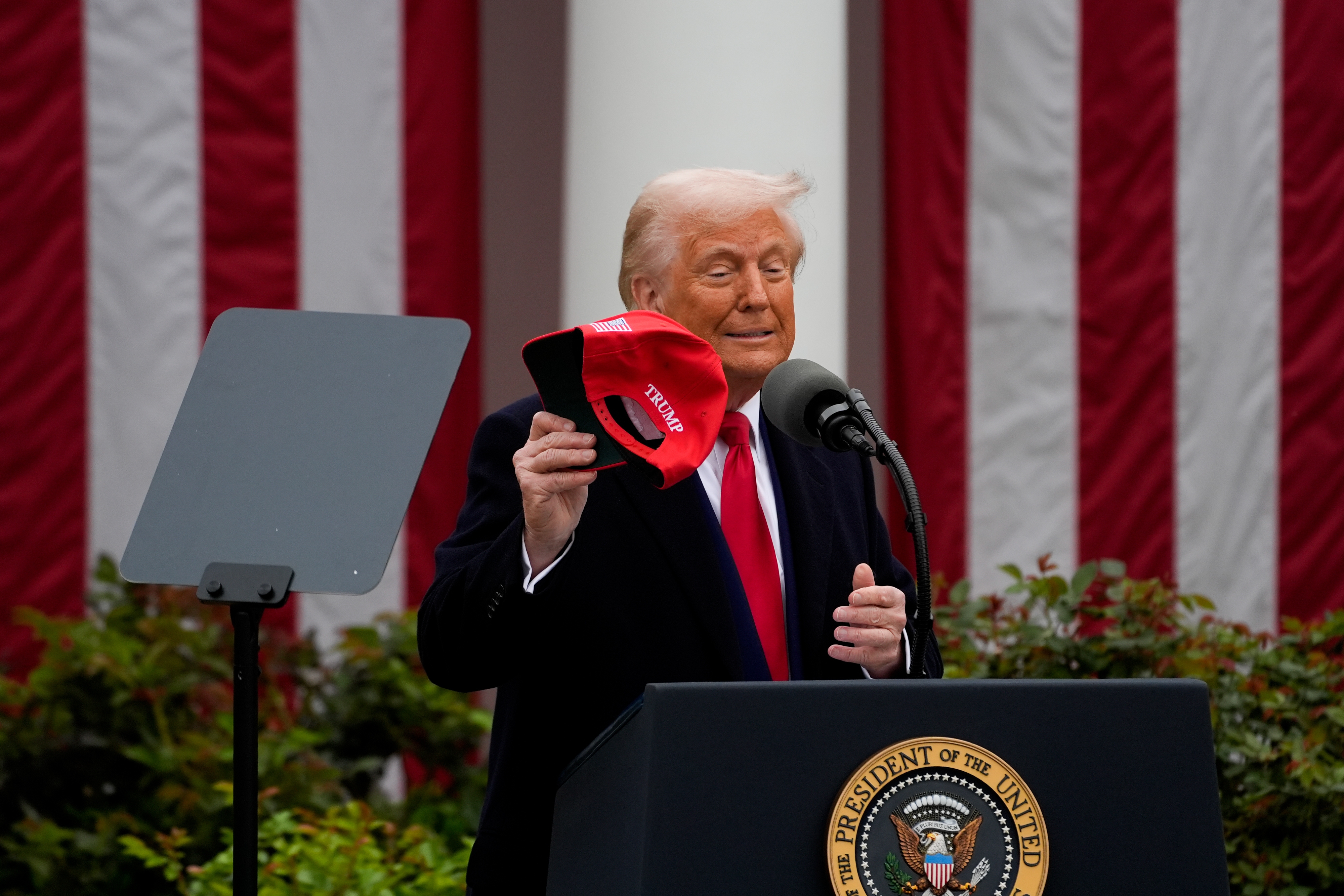 A person holding a "Make America Great Again" hat speaks at a podium in front of the U.S. presidential seal and large vertically striped backdrop