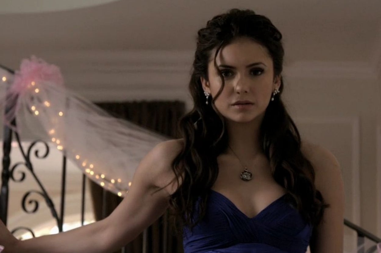 Person in elegant, strapless dress with curled hair and pendant necklace, standing indoors by a decorated railing
