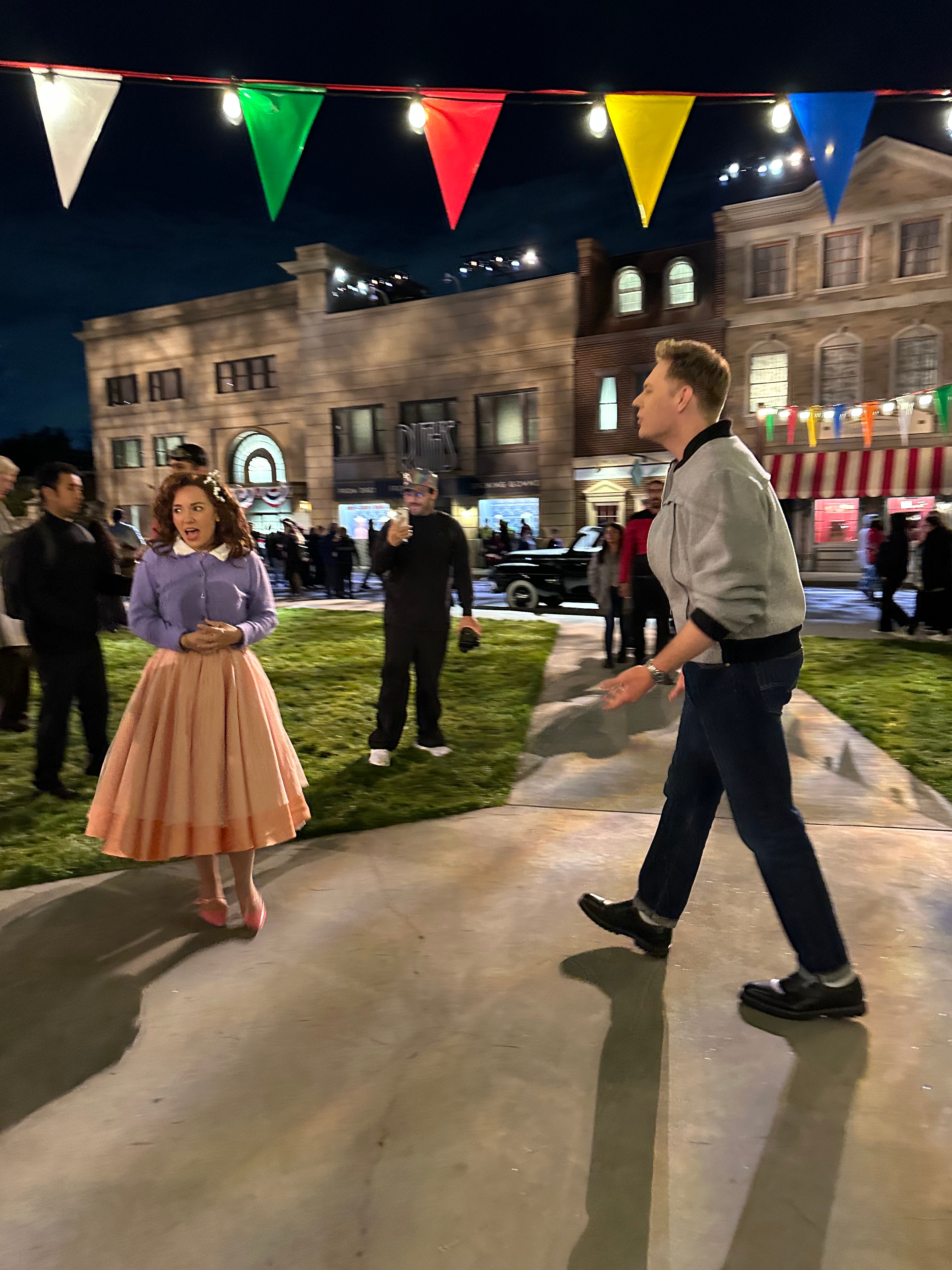 A man in casual attire and a woman in a 1950s-style outfit stand on a set resembling a retro street, with colorful flags overhead