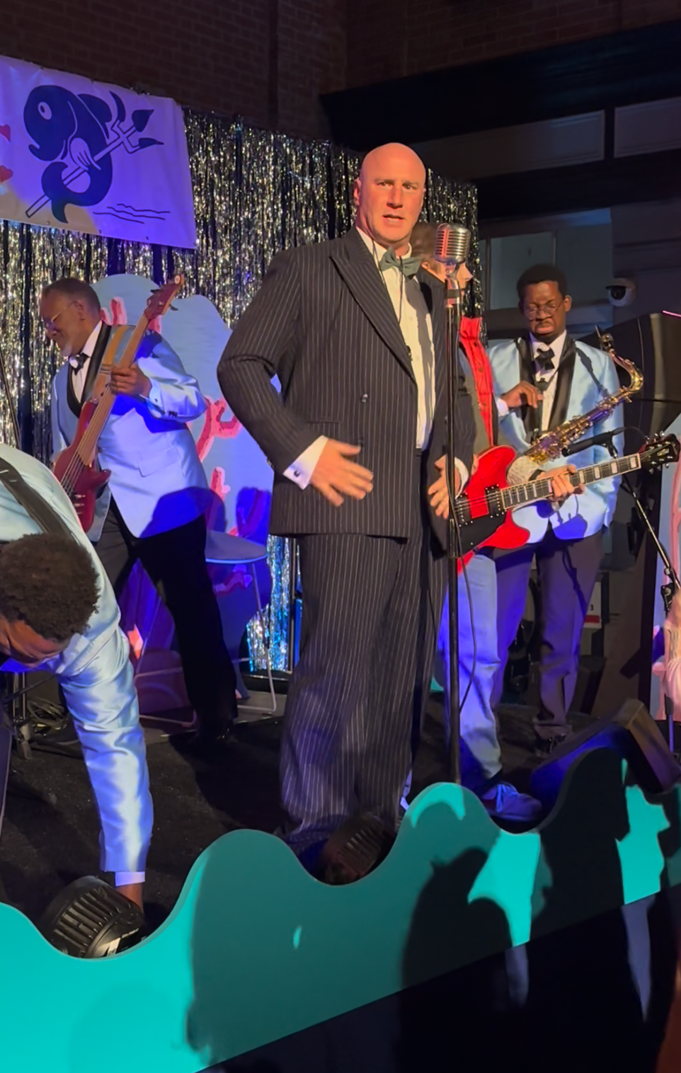 A singer in a pinstripe suit performs on stage with a vintage mic, accompanied by musicians playing guitar and saxophone, with a retro-themed backdrop