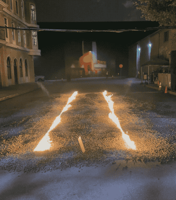 Two lines of fire on a gravel road, resembling tire tracks, in a city street at night. Buildings and a mural are visible in the background