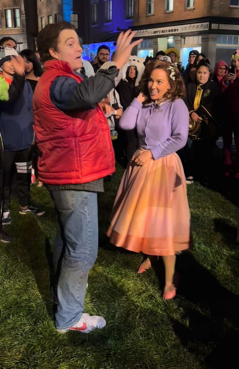 People in 1950s-inspired attire conversing outside, surrounded by onlookers. The scene appears lively and retro