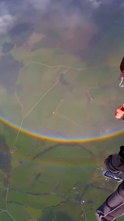 View of a skydiver mid-air above fields, with a full circular rainbow visible below them