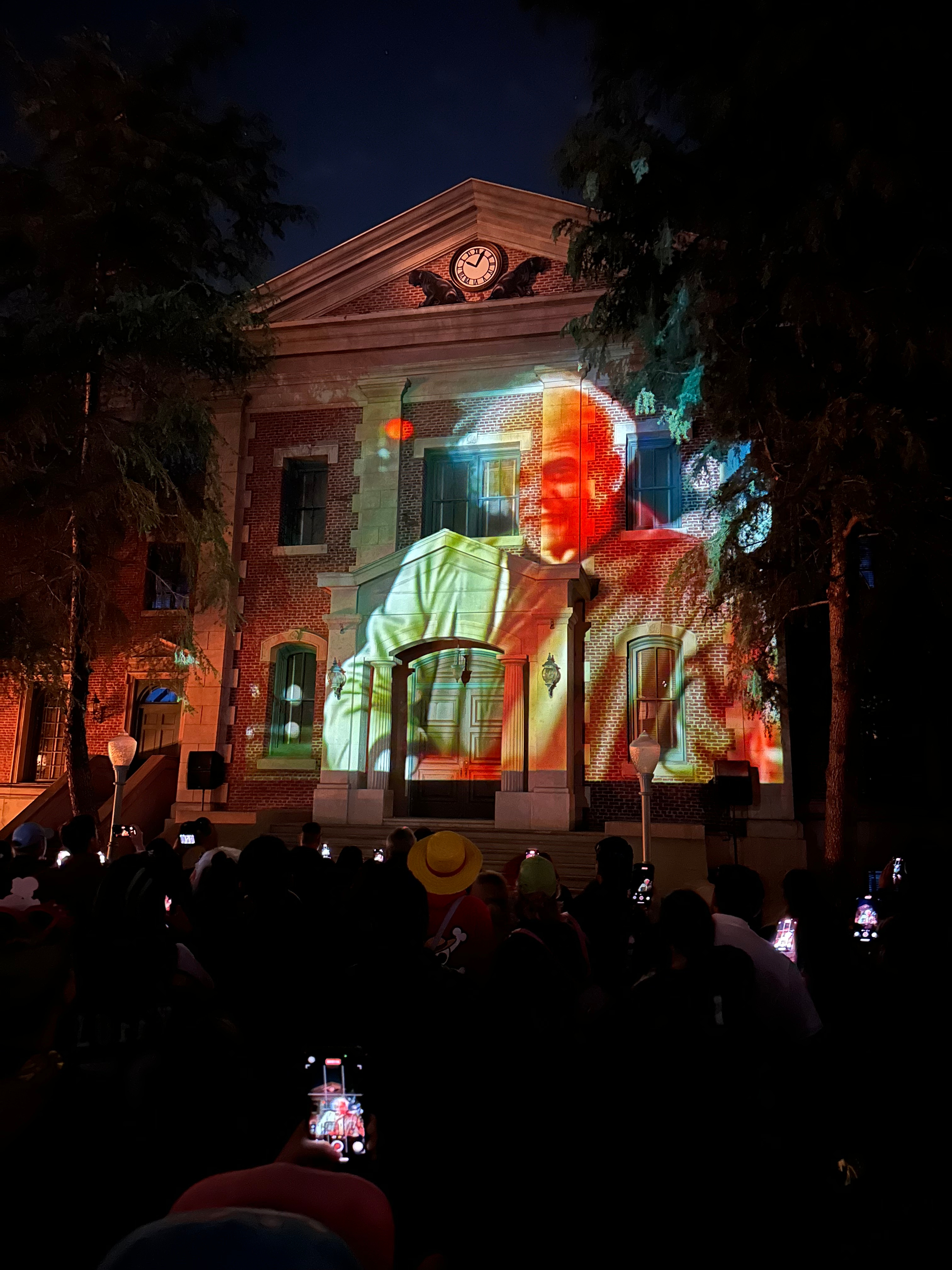 A historical building is illuminated at night with a large projection of a man speaking, viewed by a crowd holding up phones