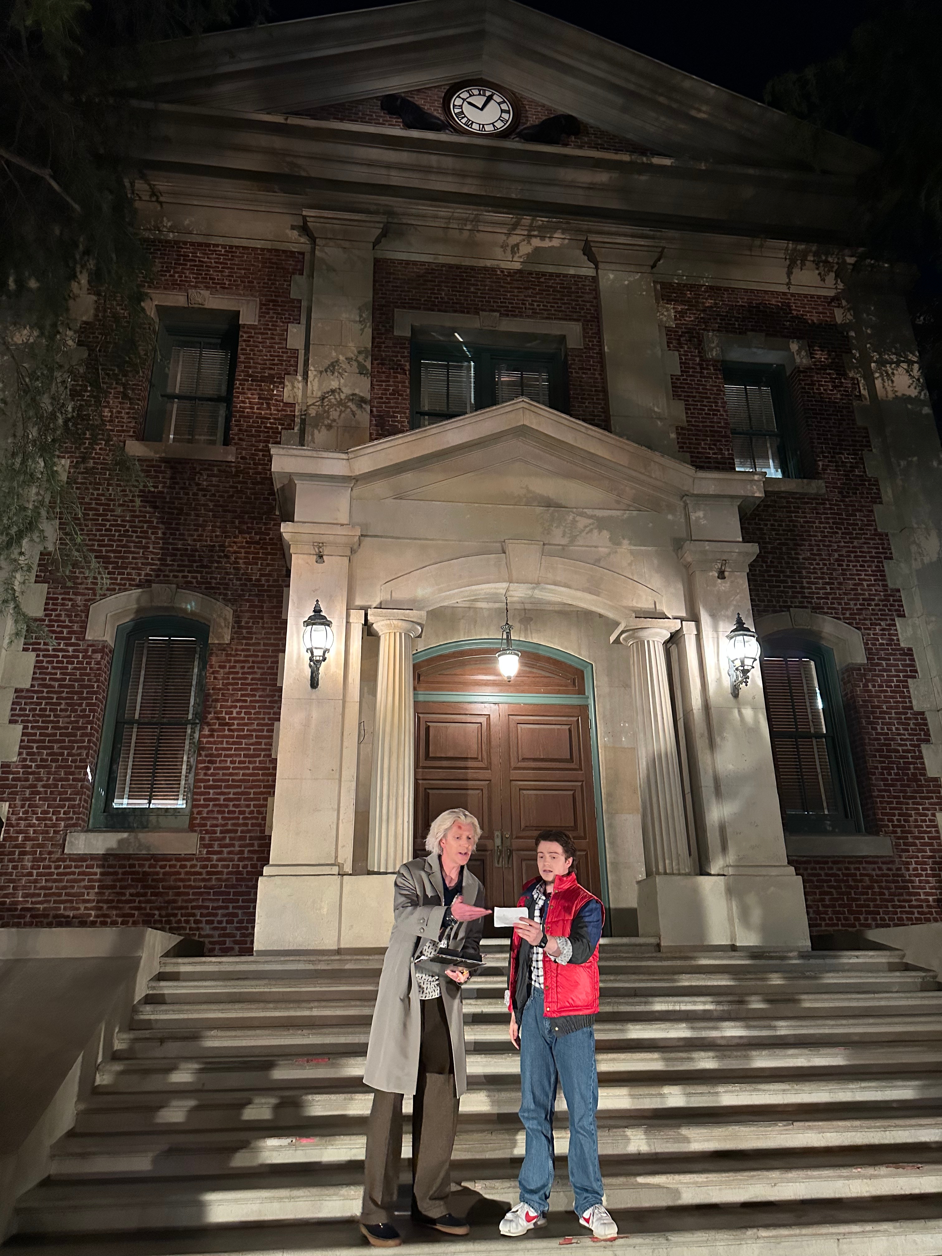 Two people in costumes stand on steps of a stately building at night, one dressed in a lab coat, the other in a puffy vest