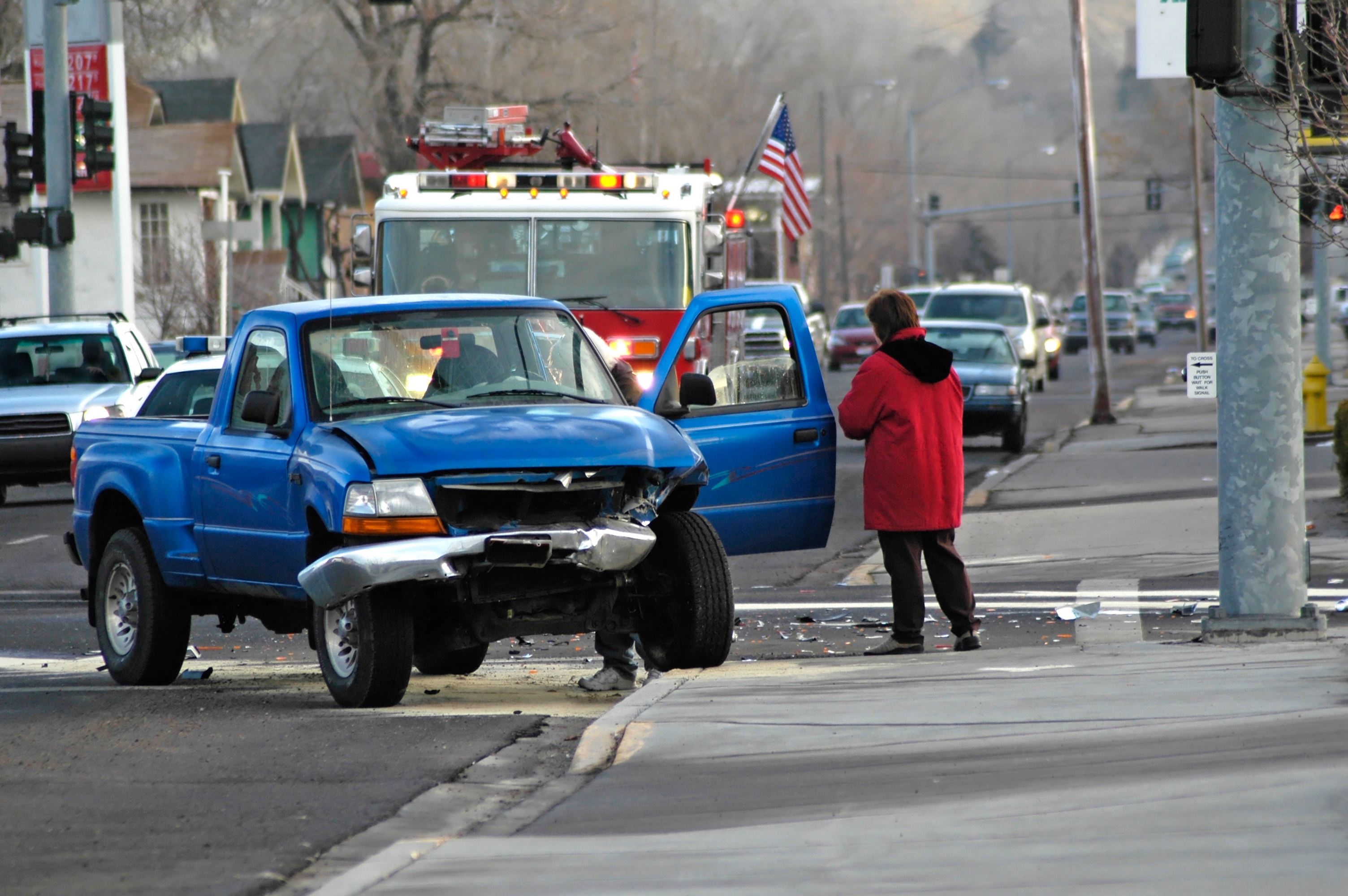 A blue pickup truck with front damage is stopped on a street. A person in a red coat stands nearby, and a fire truck is in the background