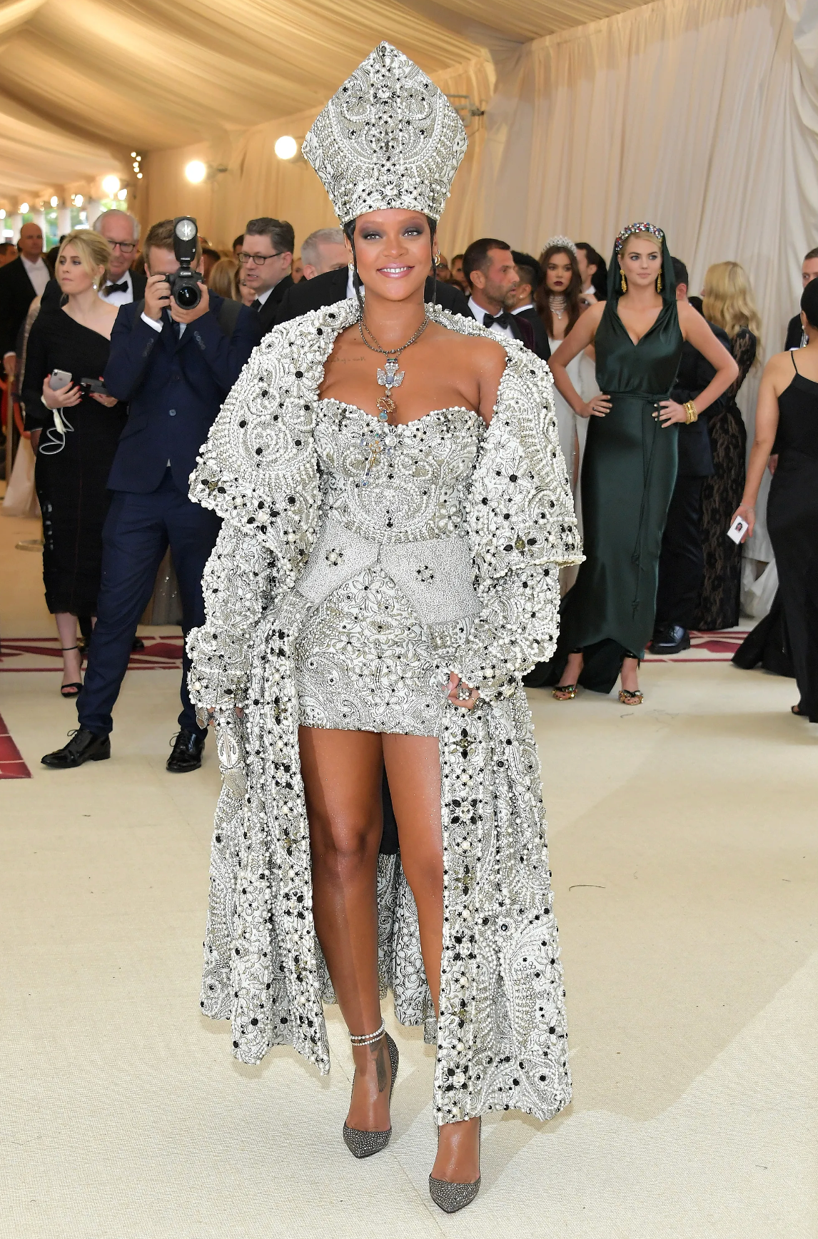 Persona con atuendo adornado de pedrería y tocado elaborado, posando en un evento de alfombra roja. Otros asistentes en segundo plano