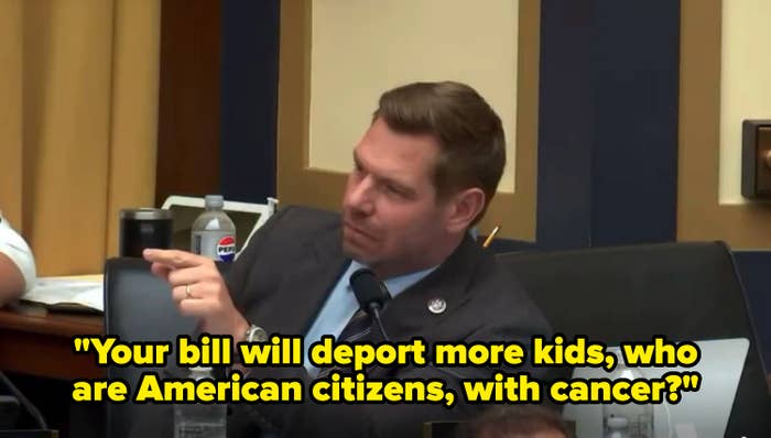 Person in a suit speaks into a microphone in a formal setting, gesturing with hand, possibly during a hearing or meeting