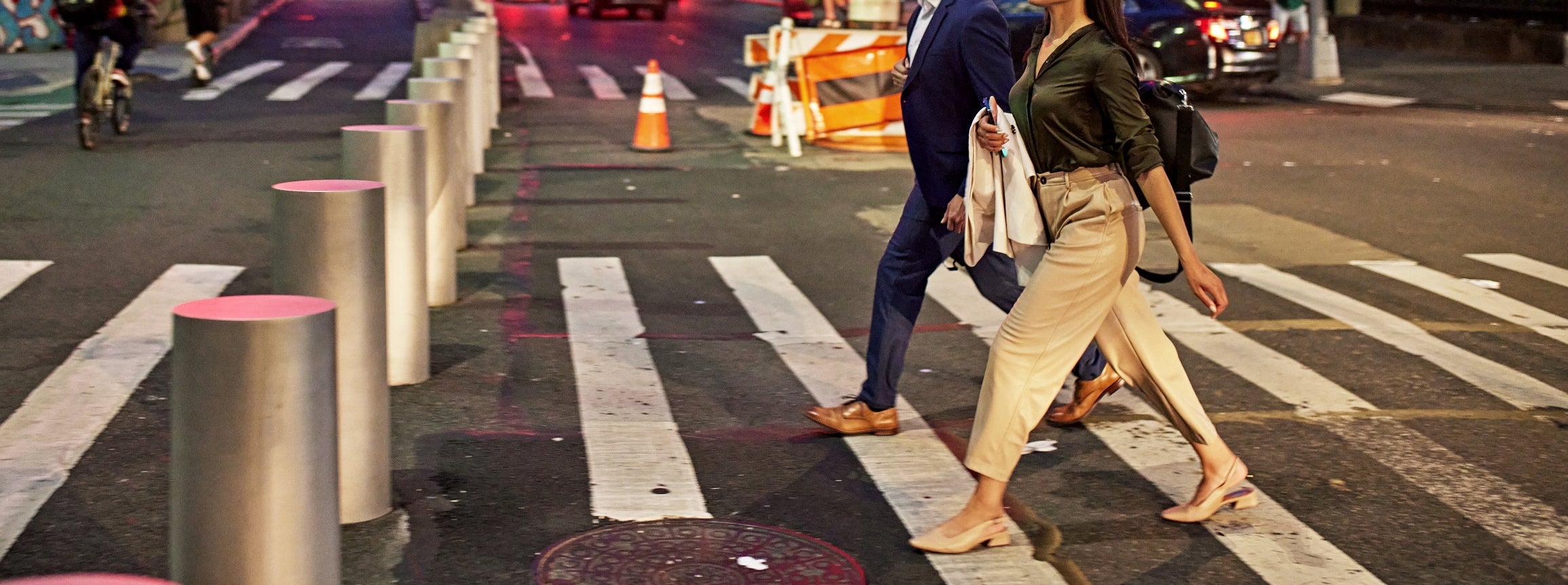 A couple in business attire crosses a busy city street at night, with traffic lights and bustling activity in the background