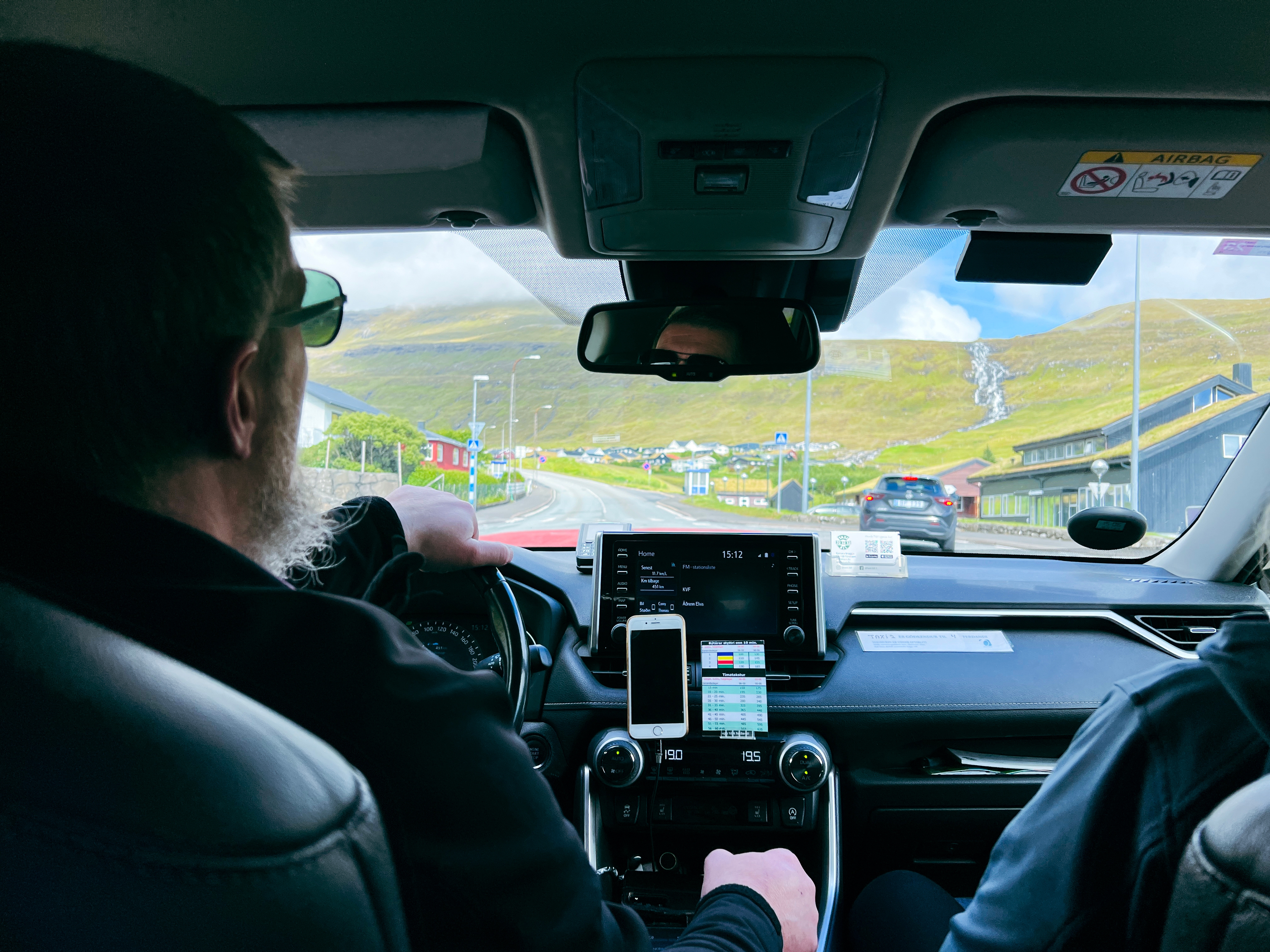 View from back seat of a car, showing two people in the front seats and a scenic road ahead with hills and buildings