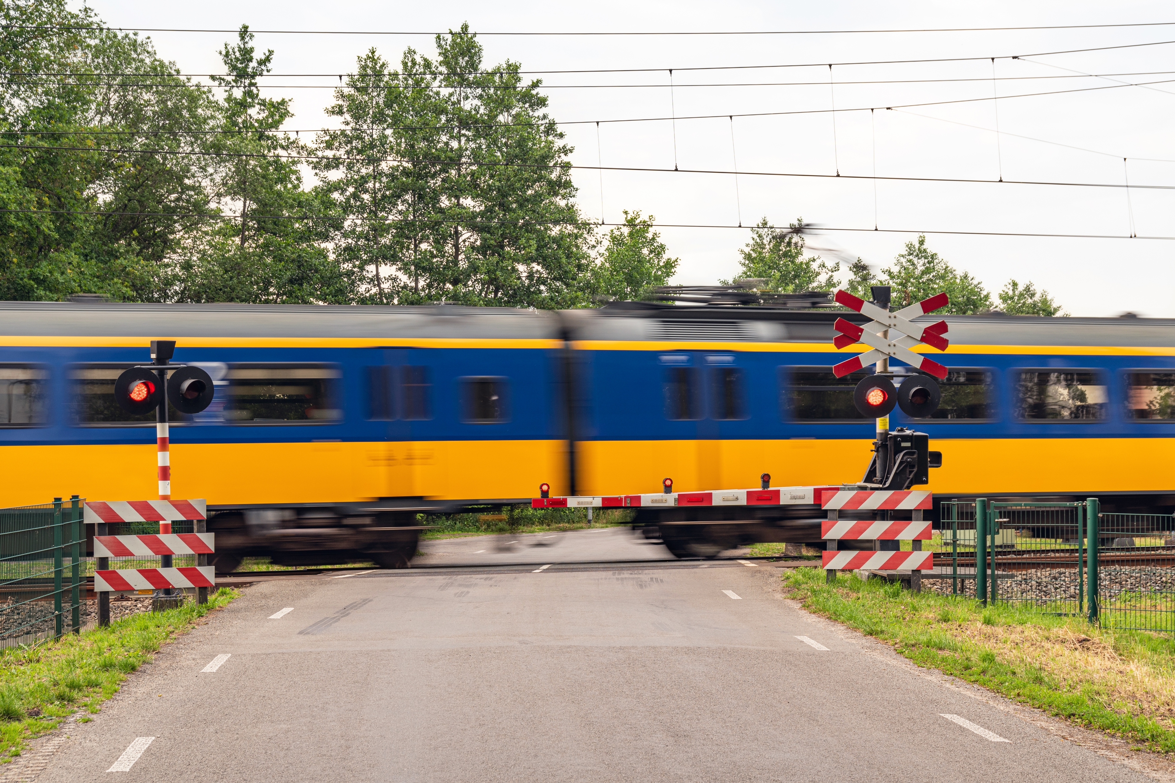 Train speeds past a level crossing with red flashing lights signaling. Trees and railway tracks are visible in the background